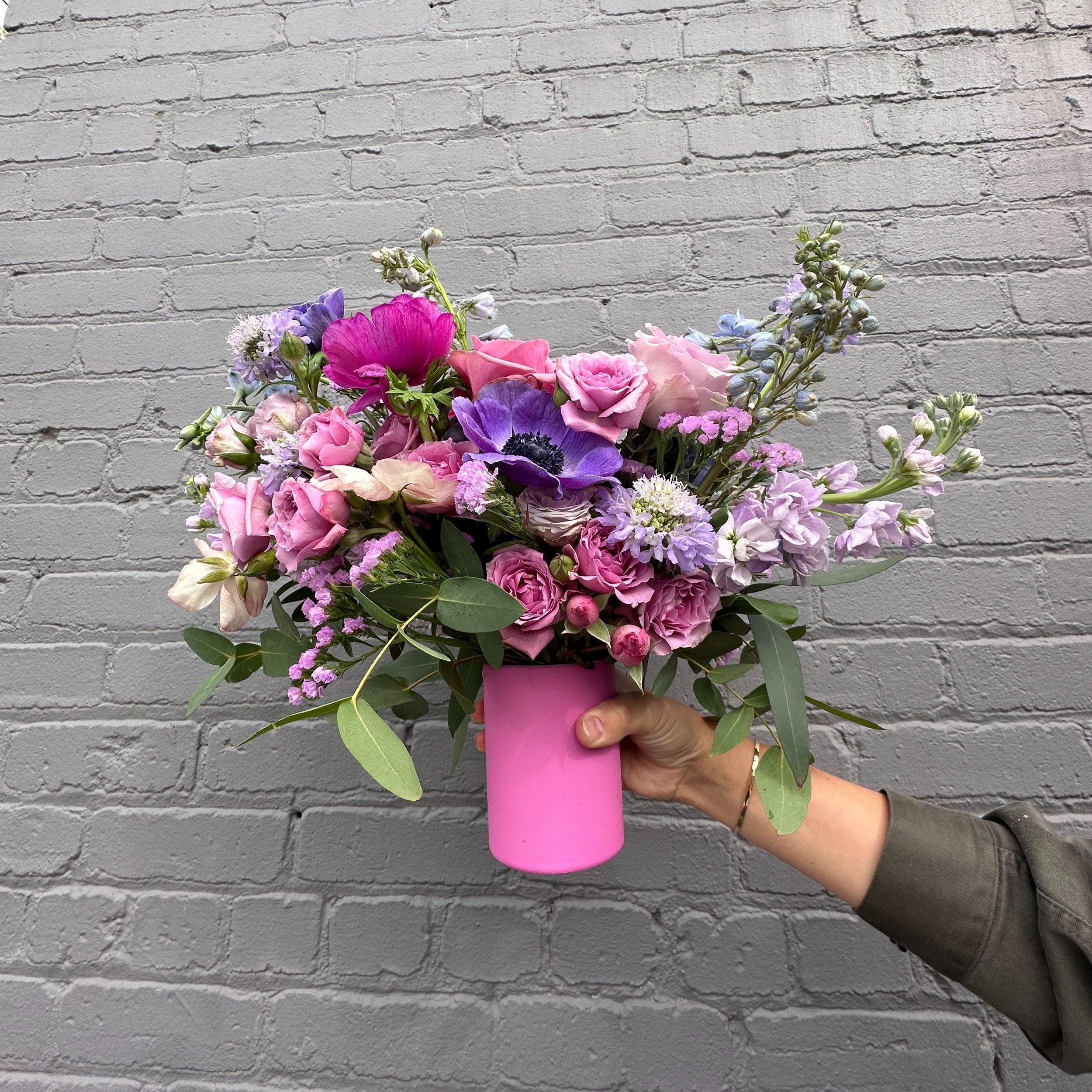Medium sized vase arrangement with pink and purple flowers being held by one hand in front of a grey brick wall