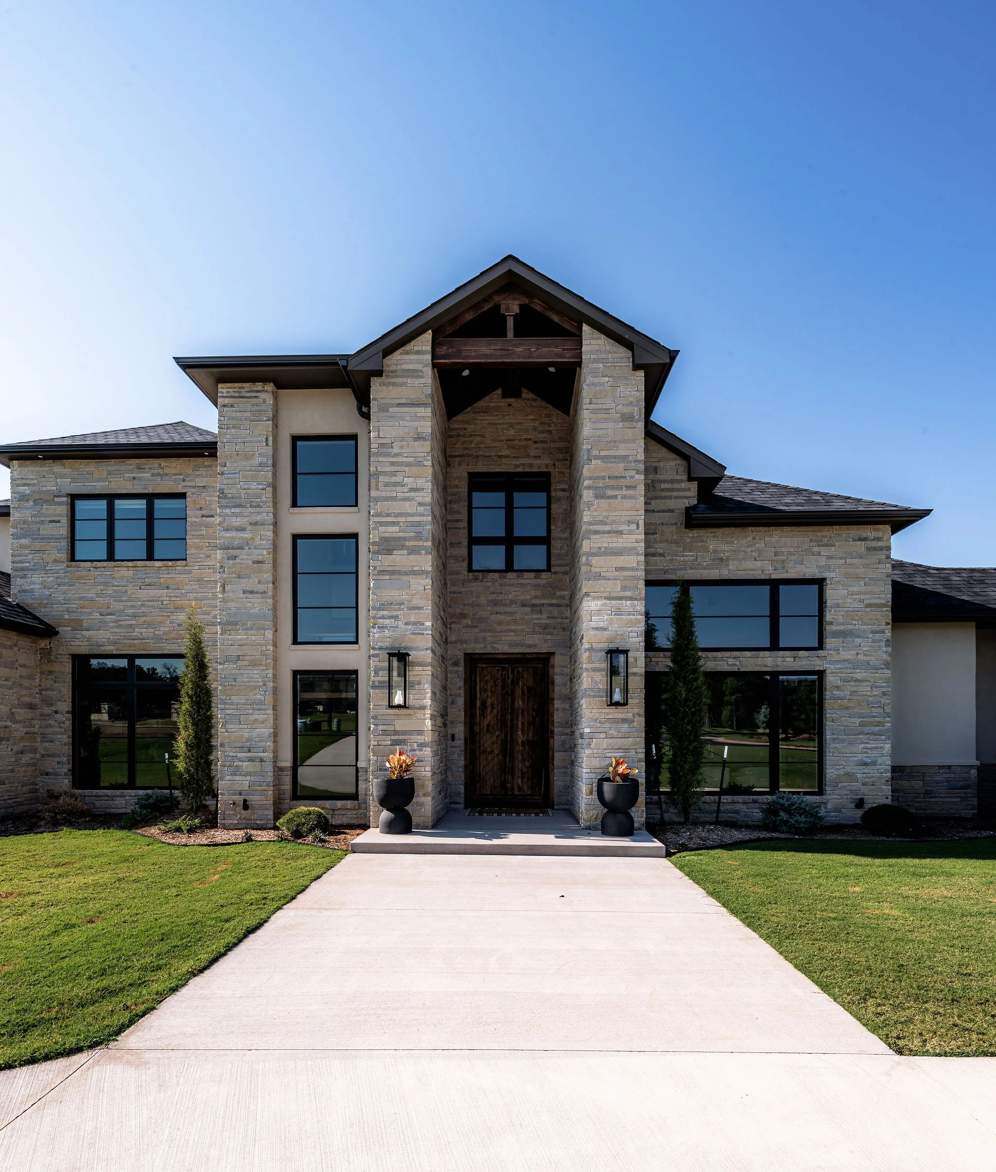 Modern house with stone exterior, large windows, and a wooden front door, surrounded by a well-maintained lawn and concrete pathway.