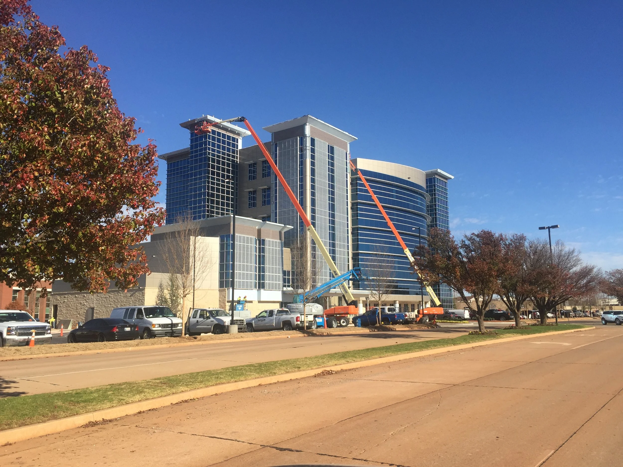 Construction work in front of a tall modern building with glass windows, using cranes and lifts, in a parking lot with cars and trees with autumn leaves.