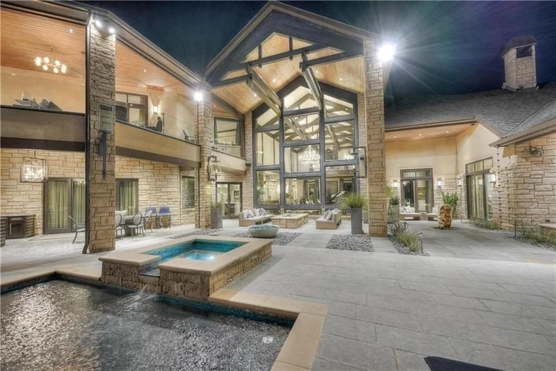 Night view of a modern house with a spacious outdoor patio, hot tub, and water feature, featuring large glass windows and a peaked roof.