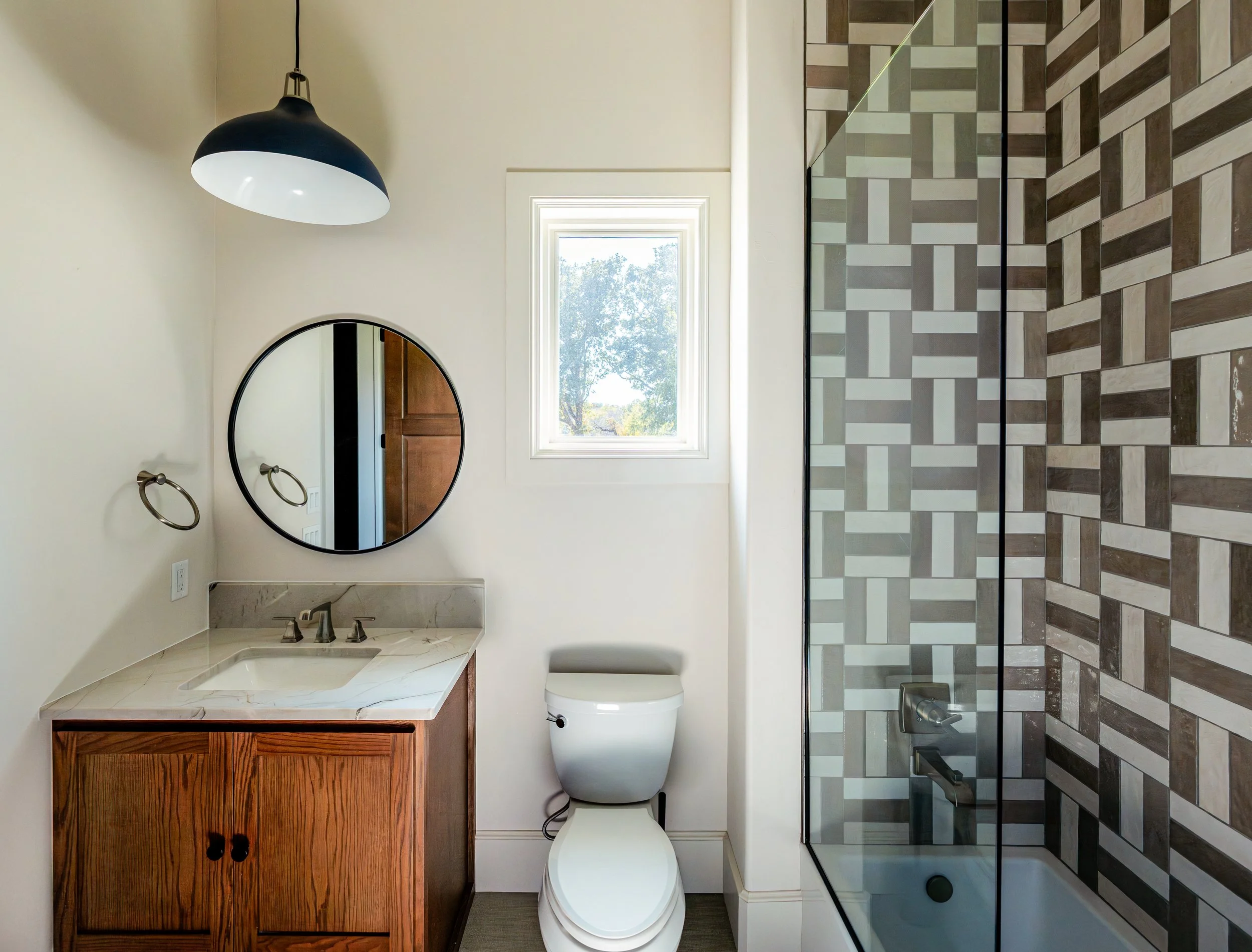 Bathroom with a hanging black and white light fixture, round mirror, wooden vanity with a marble countertop, white toilet, small window, and a bathtub with a glass shower door and decorative tile wall.