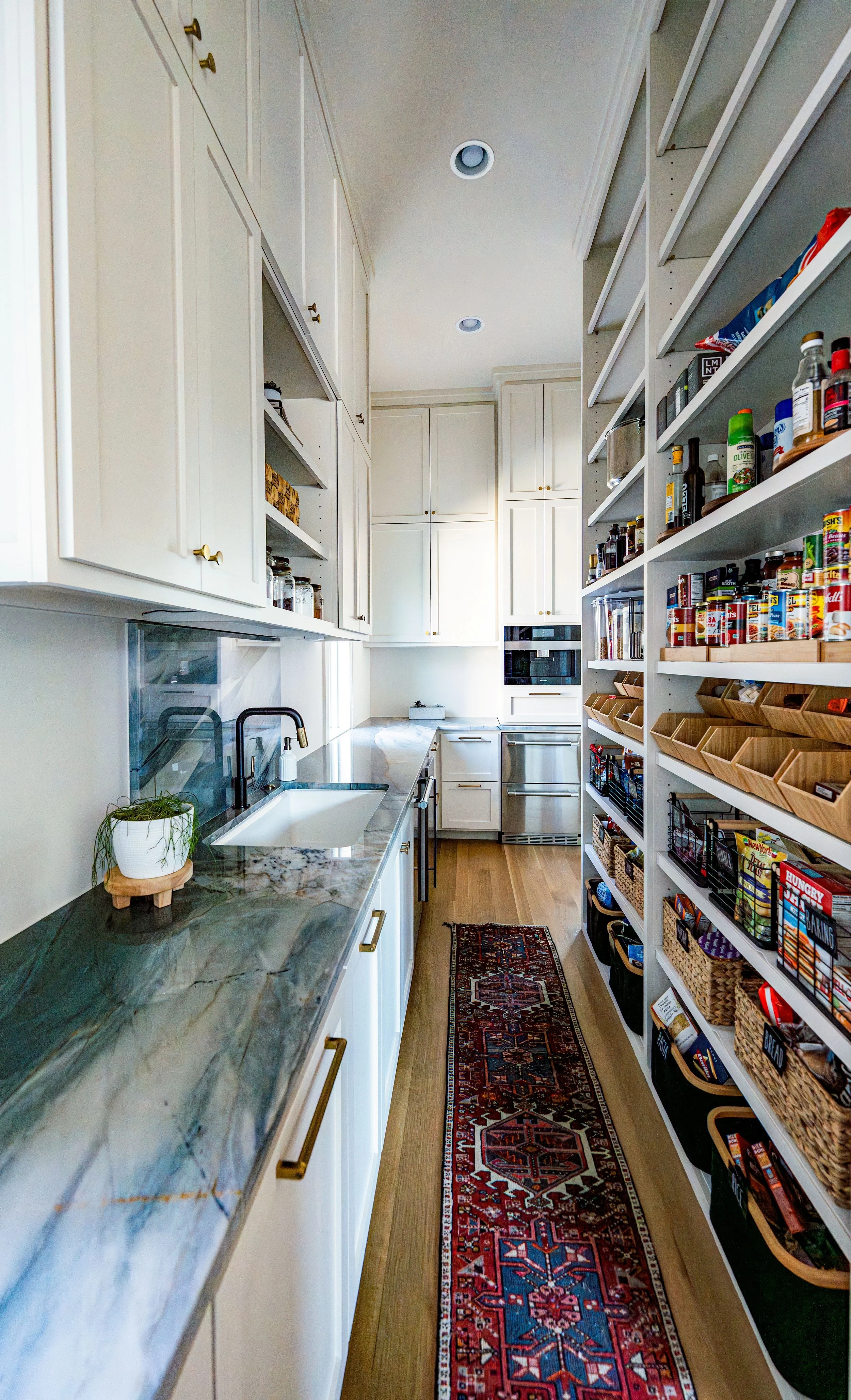 A long, narrow pantry with white cabinets, open shelves with spices and pantry items, a marble countertop, and a small rug on wooden floor, leading to built-in oven and microwave.