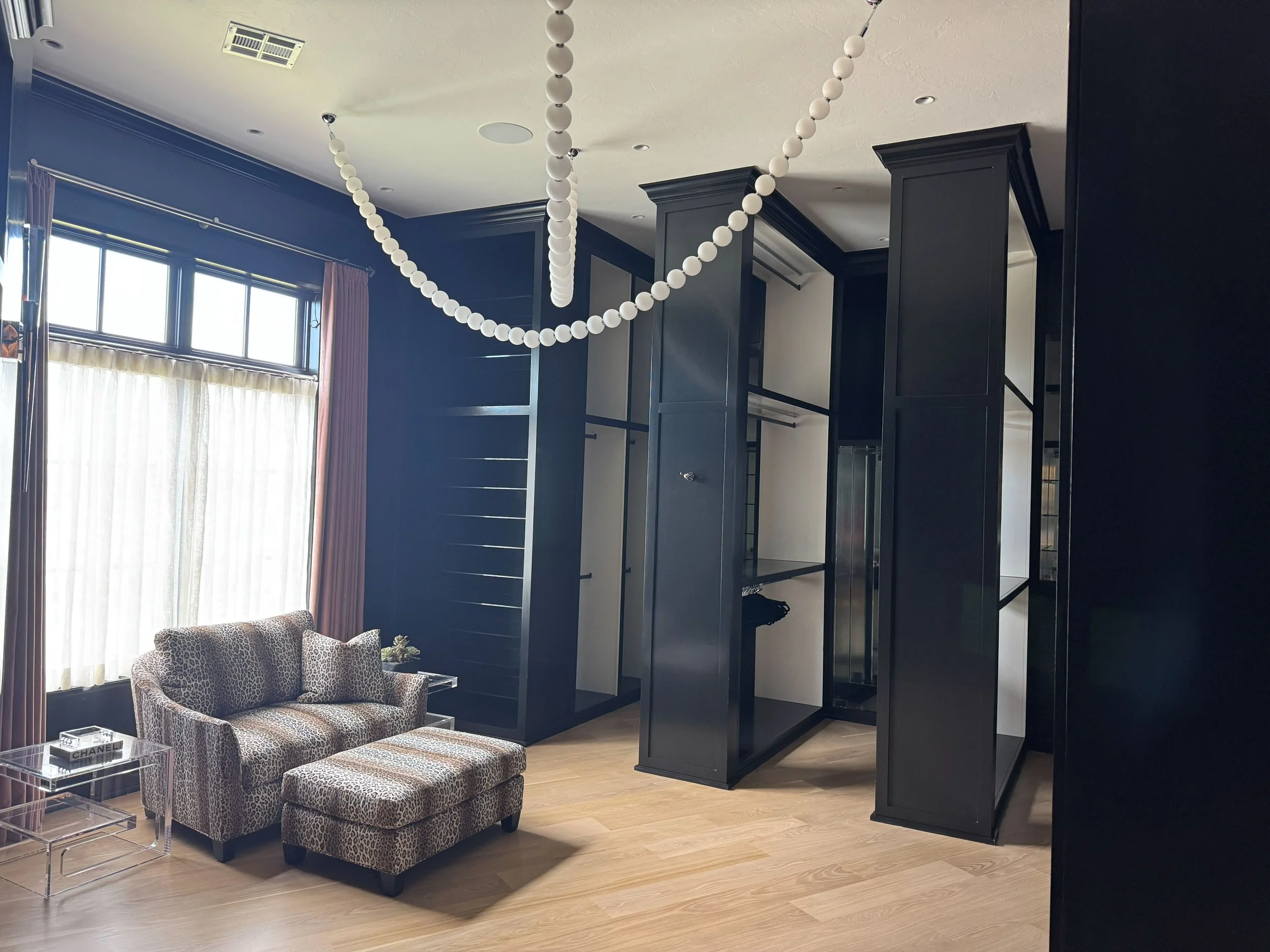 Living room with a leopard print sofa and matching ottoman, a clear acrylic side table with a Chanel box, large window with sheer and pink curtains, black modern open wardrobe, string of white decorative balls hanging from the ceiling.
