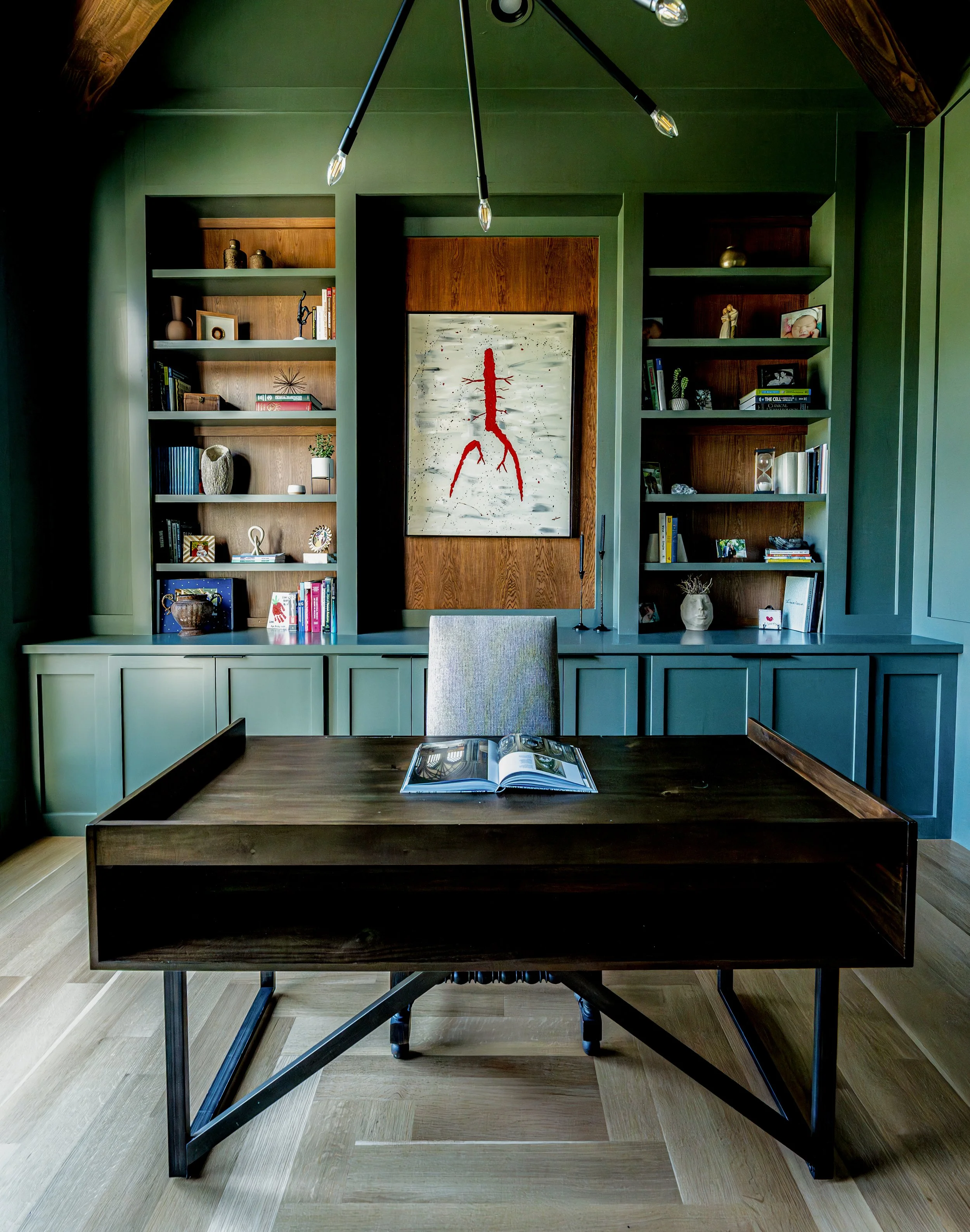 A modern office room with green walls, built-in bookshelves filled with various books and decor, a wooden desk, and a gray upholstered chair. There is a large abstract painting with red and black accents on the wall and a ceiling light fixture with e