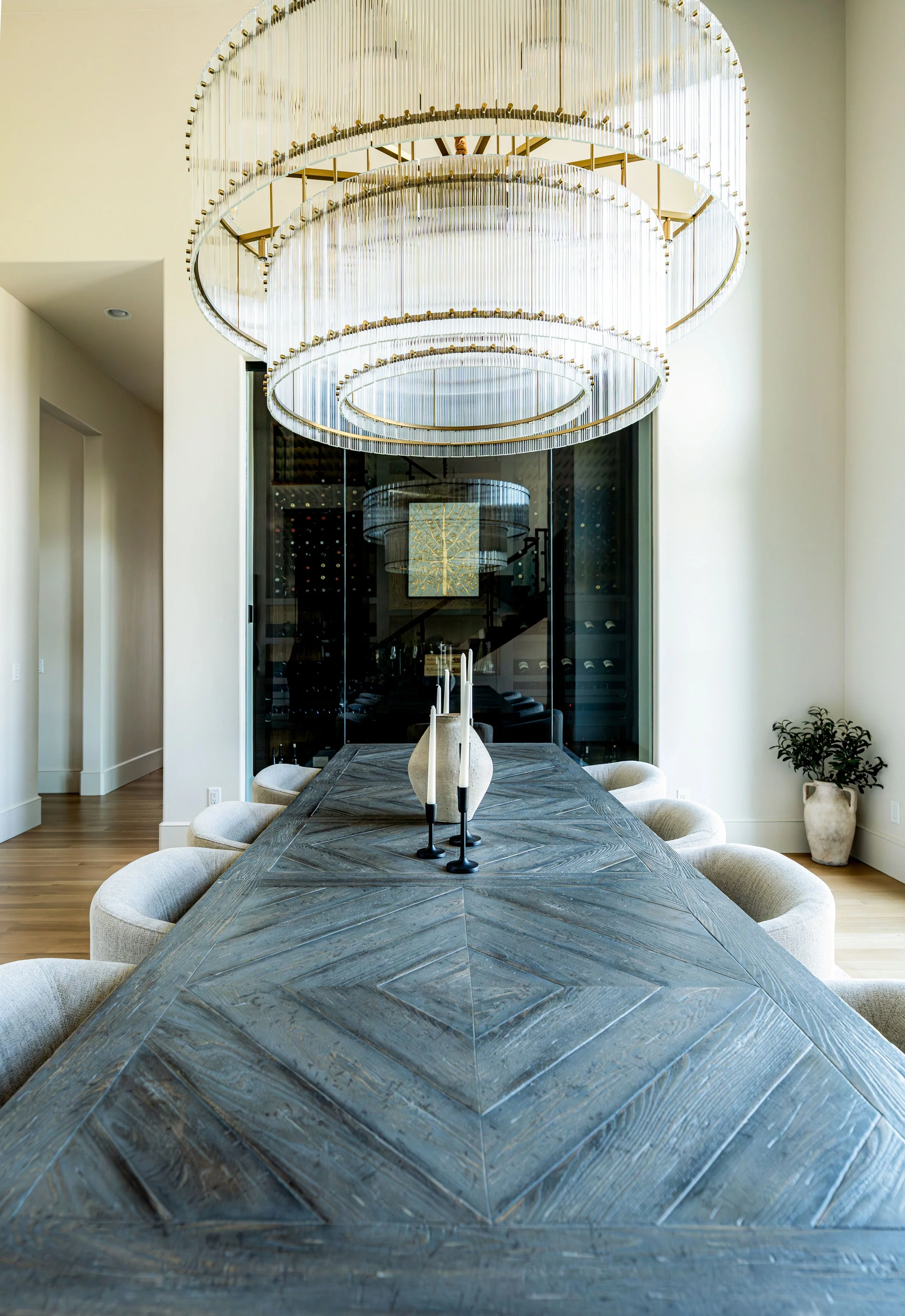 Modern dining room with a long, textured wooden table, six light beige upholstered chairs, and a large, elegant chandelier with three circular tiers hanging from the ceiling. A glass wall or door with a wine cellar behind reflects the room, and a pot