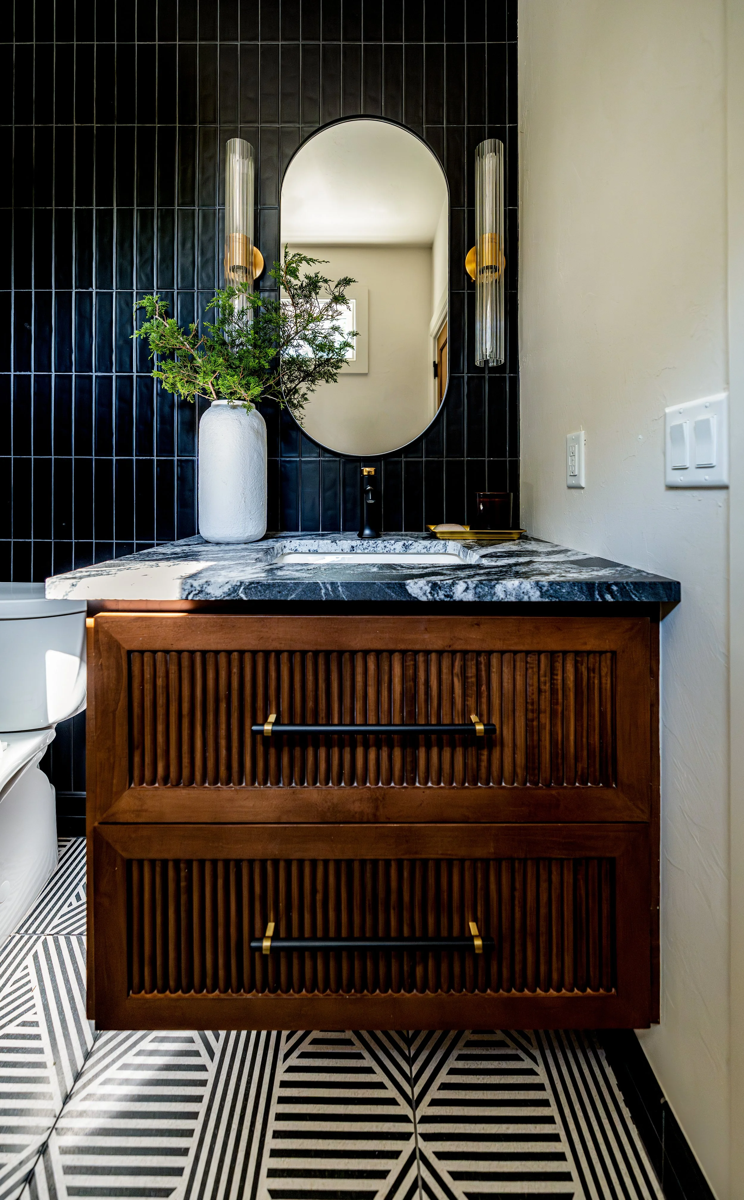 Modern bathroom vanity with a marble countertop, black hardware, and a wooden cabinet with vertical slats, a vessel vase with greenery, and a mirror flanked by two vertical light fixtures.