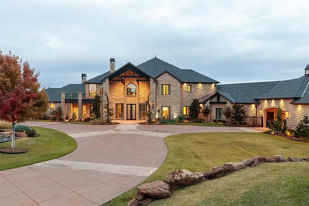 Large, elegant house with stone exterior and multiple peaked roofs, surrounded by a well-maintained lawn and trees, with soft interior lighting visible through the windows during dusk.