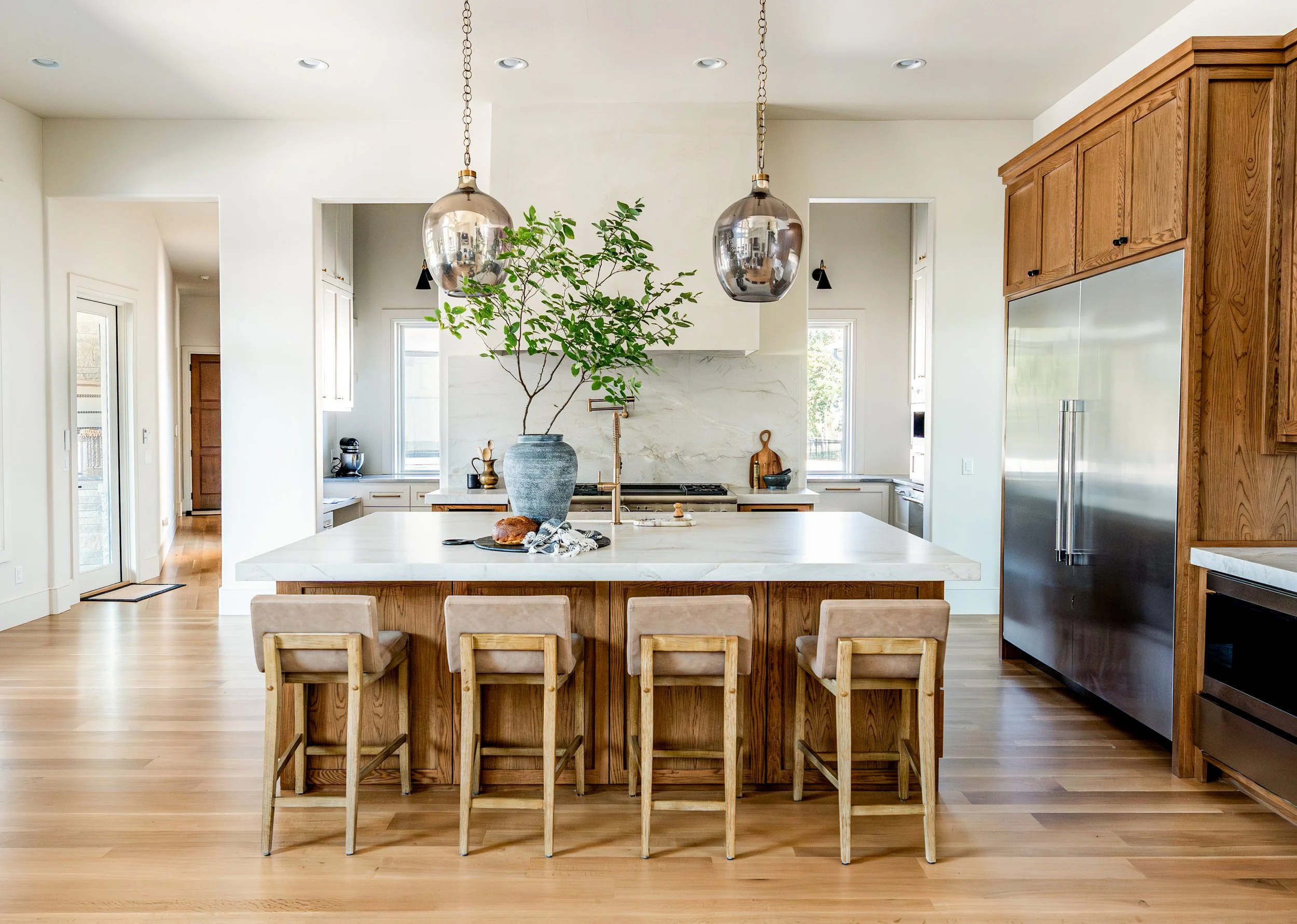 Modern kitchen with white marble island, wooden cabinetry, stainless steel refrigerator, pendant lights, and a large vase with green branches on the island.