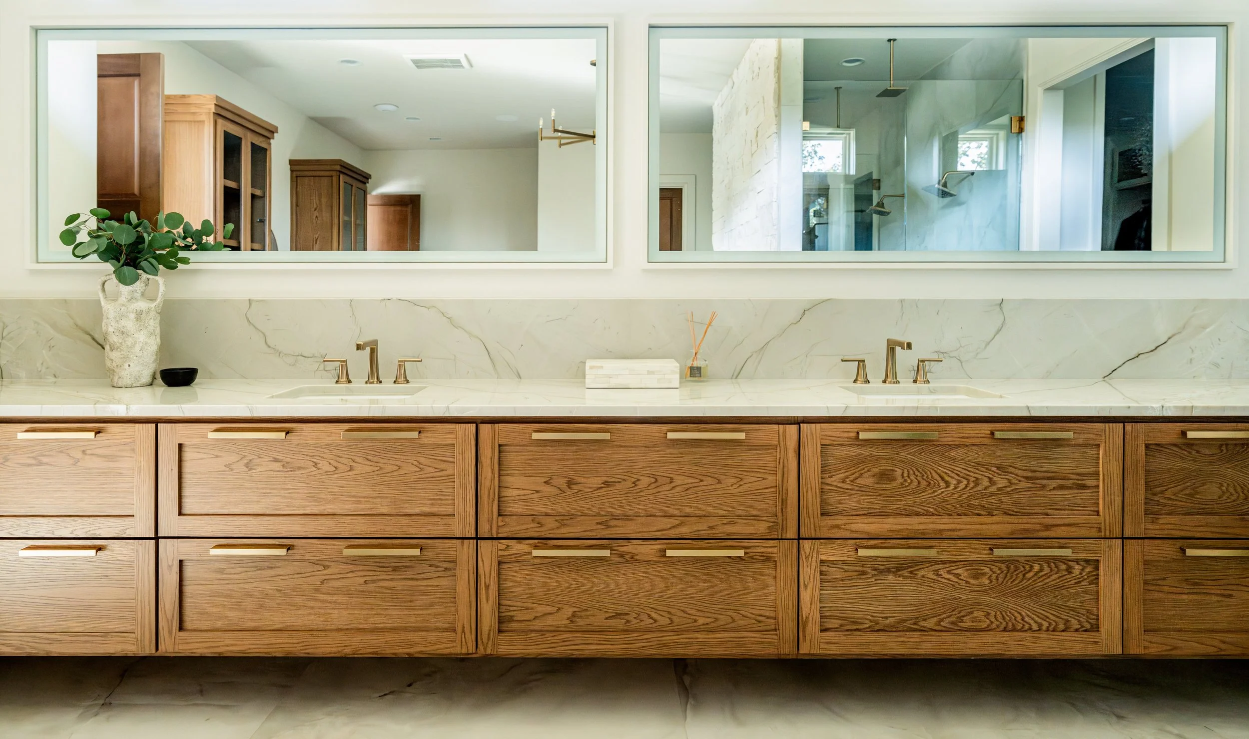 Modern bathroom vanity with wooden cabinets, marble countertop, two sinks, and a large mirror above. Decor includes a potted plant, a small black bowl, and a reed diffuser.