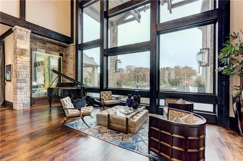 Living room with large floor-to-ceiling windows, a black grand piano, beige and patterned armchairs, a glass coffee table, decorative vases, wooden flooring, and a tall plant.
