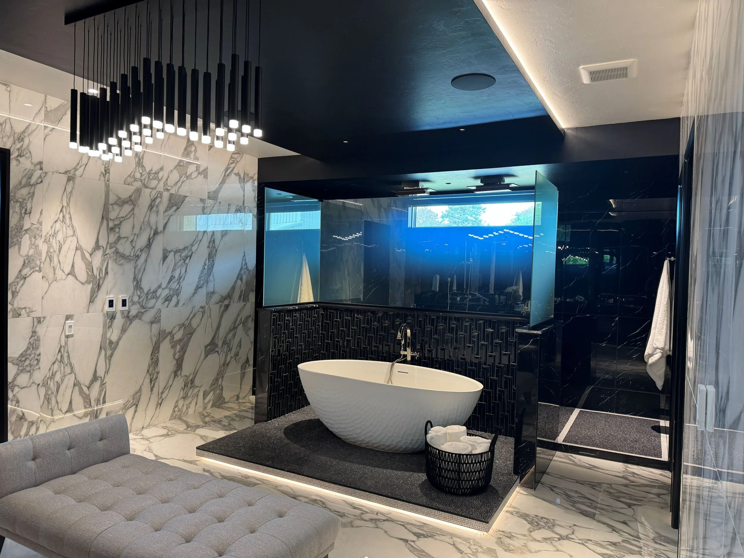 Modern bathroom with white bathtub, black accent wall, marble floors and walls, gray seating, and a glass-enclosed shower.