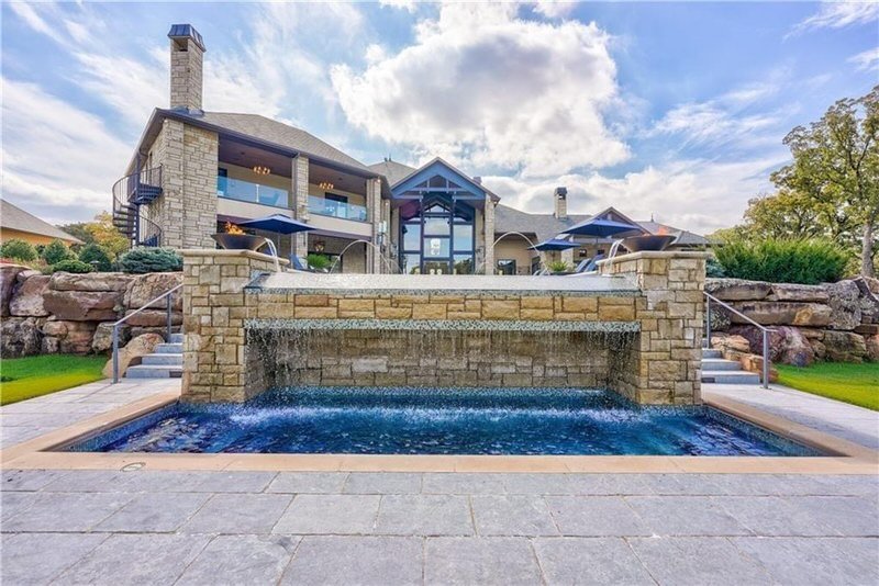 Luxury house with a waterfall feature in the backyard, stone steps leading up to a patio with umbrellas, and a glass entrance door, trees and a partly cloudy sky in the background.