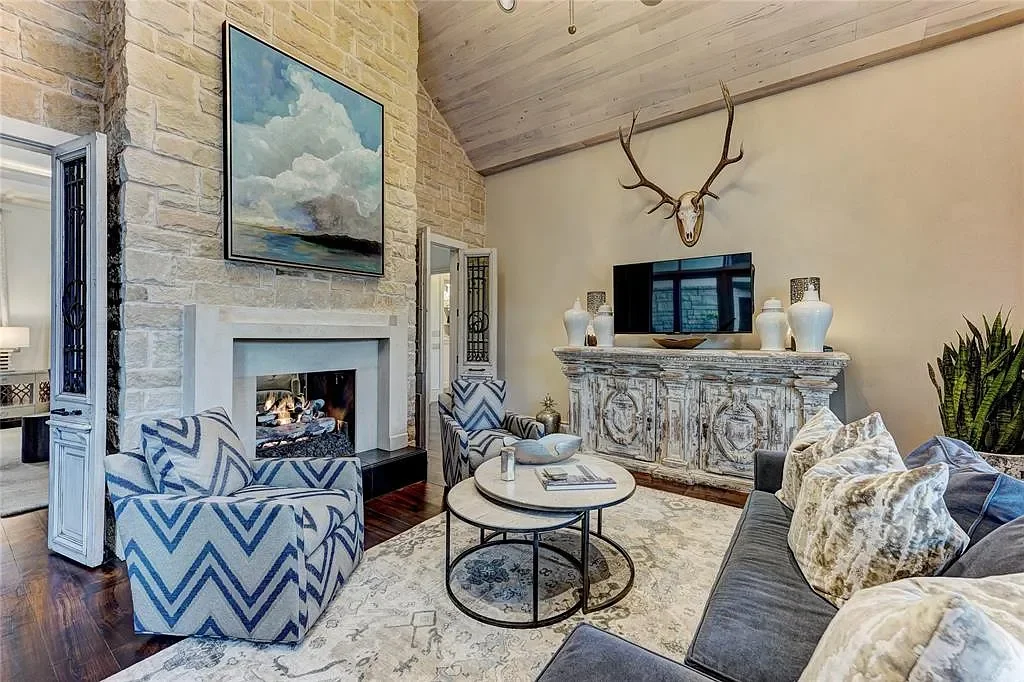 Living room with stone fireplace, blue and white chevron armchairs, gray sofa with patterned pillows, rustic wooden cabinet, mounted deer skull with antlers, flat-screen TV, and decorative vases.