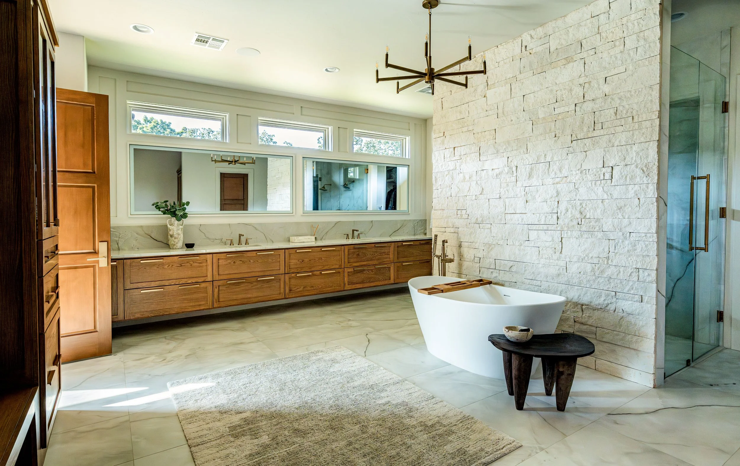Modern bathroom with a freestanding bathtub, white stone accent wall, large window with three transom windows, wooden vanity with double sinks, large mirror, and a dark wooden side table.