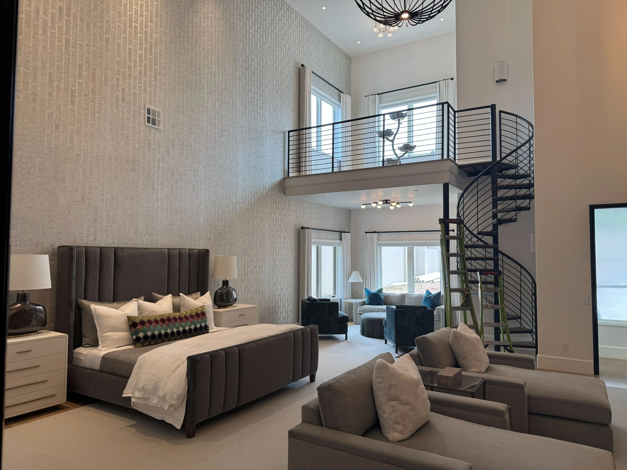 Spacious living room with a bedroom in a modern home, featuring a high ceiling, large windows, a black spiral staircase, seating area with armchairs, a bed with lamps on nightstands, and a wall with textured wallpaper.