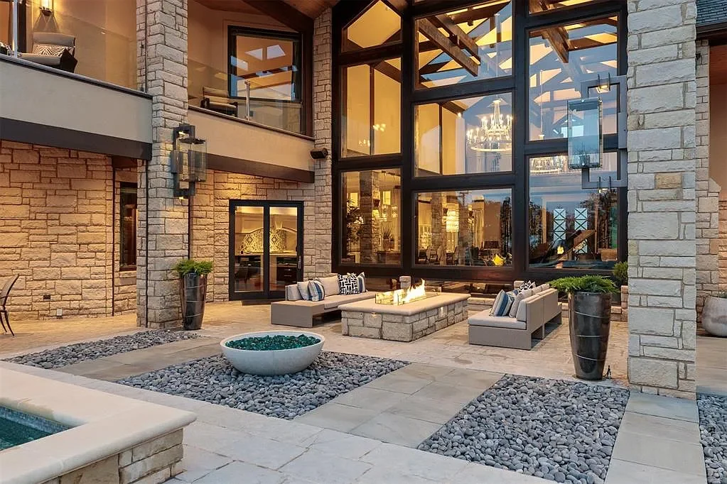 Modern outdoor patio with seating area, fire pit, potted plants, and large glass windows reflecting interior chandelier, set against a stone house exterior.