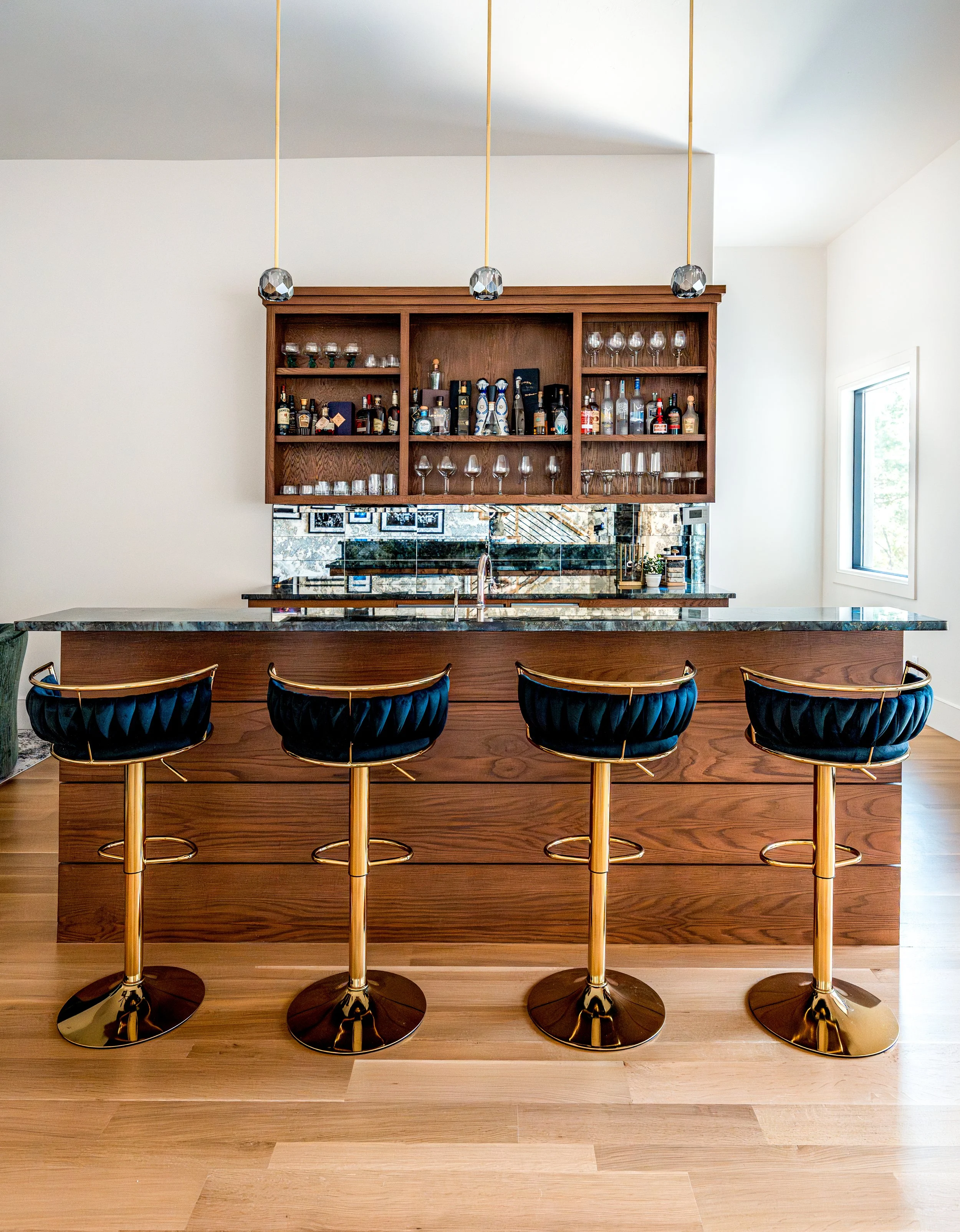 Modern home bar with a wooden counter, four black and gold bar stools, and a wooden shelf with various bottles and glasses, near a window with natural light.