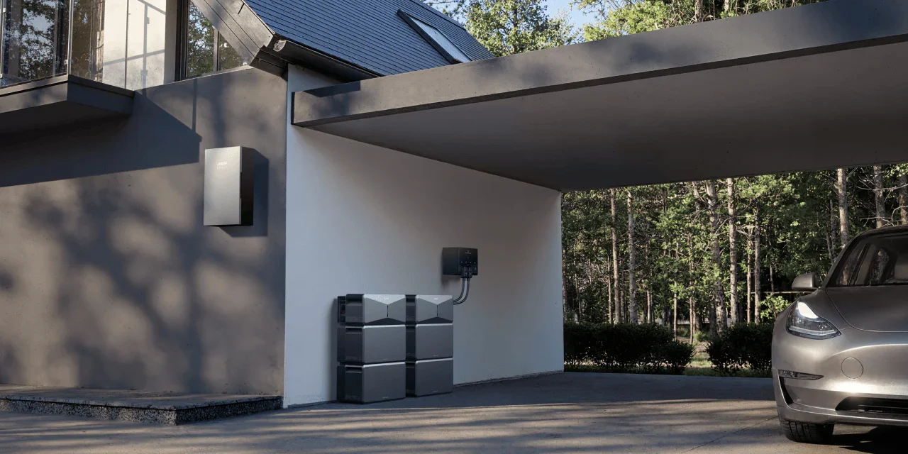 Modern house exterior with a gray and white facade, mounted air conditioning unit, a stack of gray composters, and a silver car parked in the driveway near a wooded area.