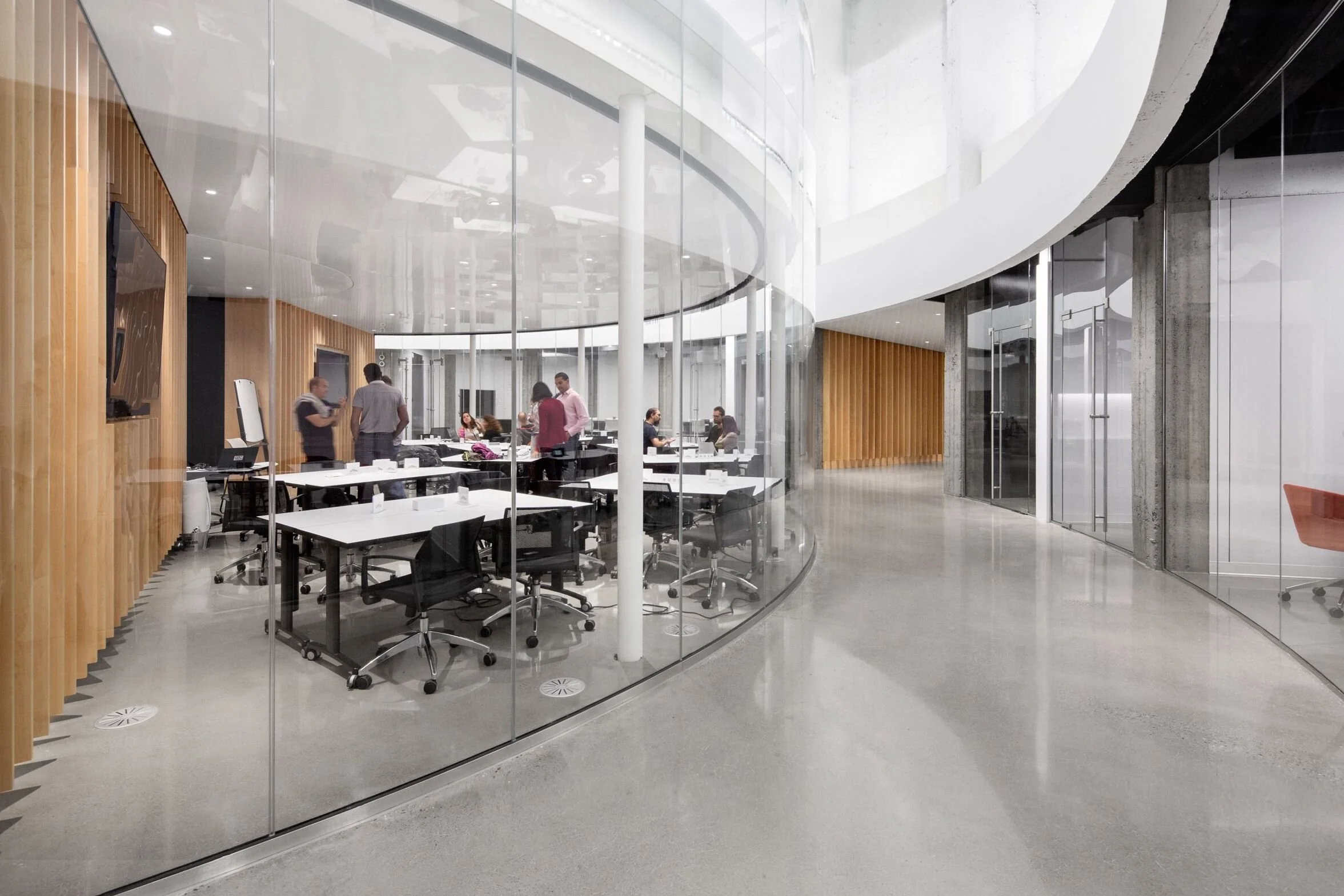 Couloir d'un espace de bureau moderne aux couleurs grises et blanches où les sections du bureau sont séparées par des murs en verre. À l'intérieur d'une section sur la gauche, environ 12 personnes discutent et échangent des idées.