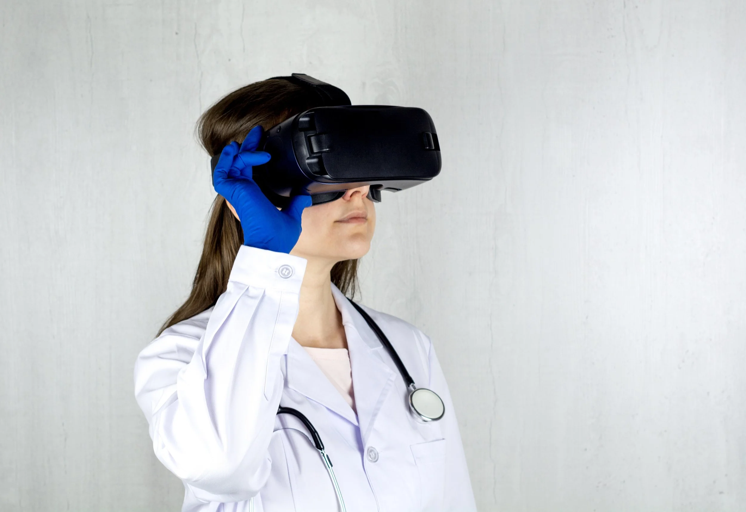Un médecin portant une blouse de laboratoire blanche et des gants de chirurgie bleus essayant un casque VR tout en regardant vers le haut devant un mur blanc.