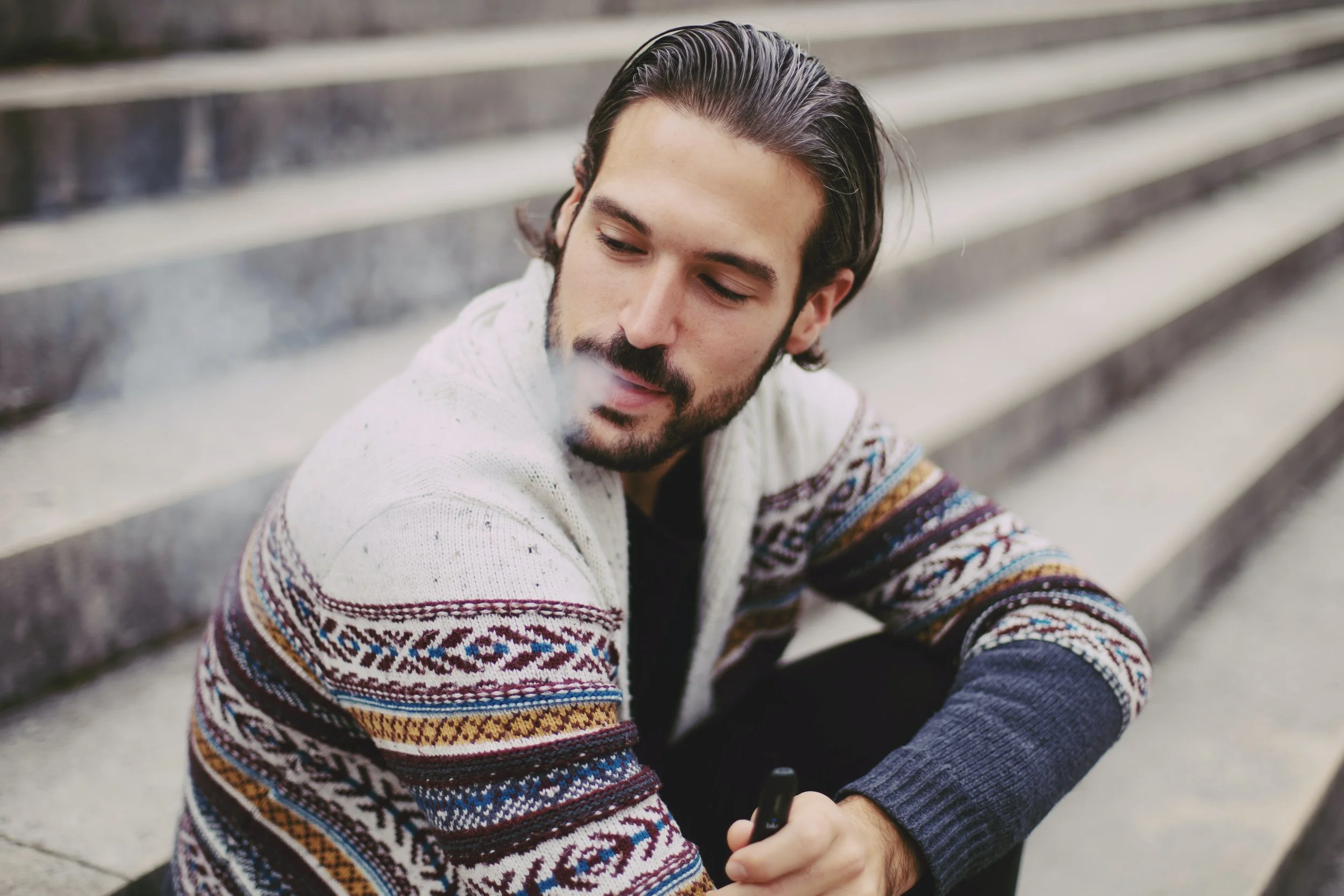 Un homme aux cheveux longs et raides poussés vers l'arrière, vêtu d'un pantalon noir et d'un cardigan coloré, est assis sur un escalier tout en fumant un stylo vape ou une cigarette électronique tout en regardant vers le bas à sa droite.