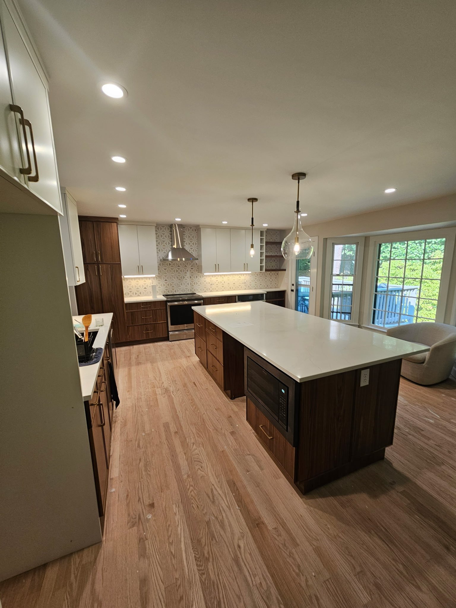 Modern kitchen with wooden floors, white and wood cabinetry, a large island with a white countertop, and two pendant lights hanging over the island. Large windows and sliding doors let in natural light.