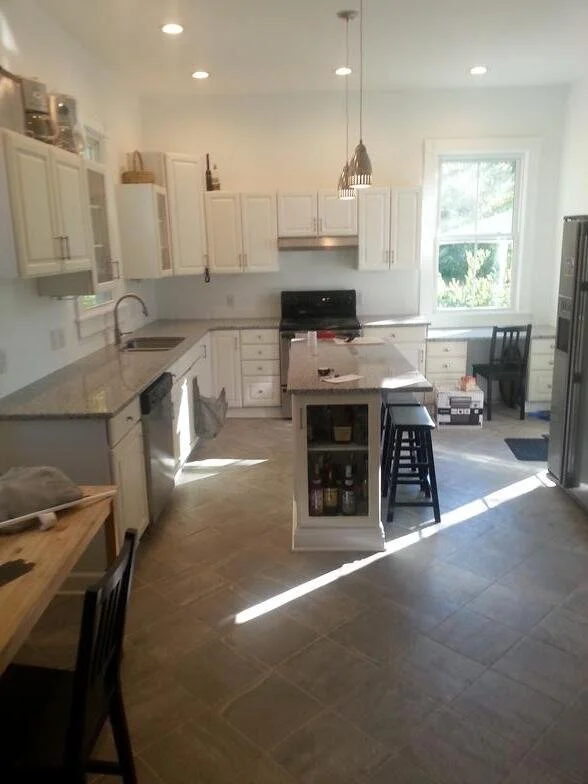 Bright white kitchen with granite countertops, white cabinets, and stainless steel appliances. An island with a small open shelf, black stools, a window letting in natural light, and some chairs and boxes in the background.