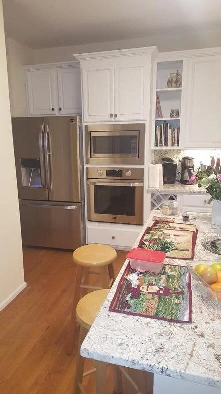 Kitchen with white cabinets, stainless steel refrigerator, microwave, oven, and a breakfast bar with two wooden stools. Countertop has holiday-themed place mats, a pink container, and fruits. Shelves with books and decorative items are visible.