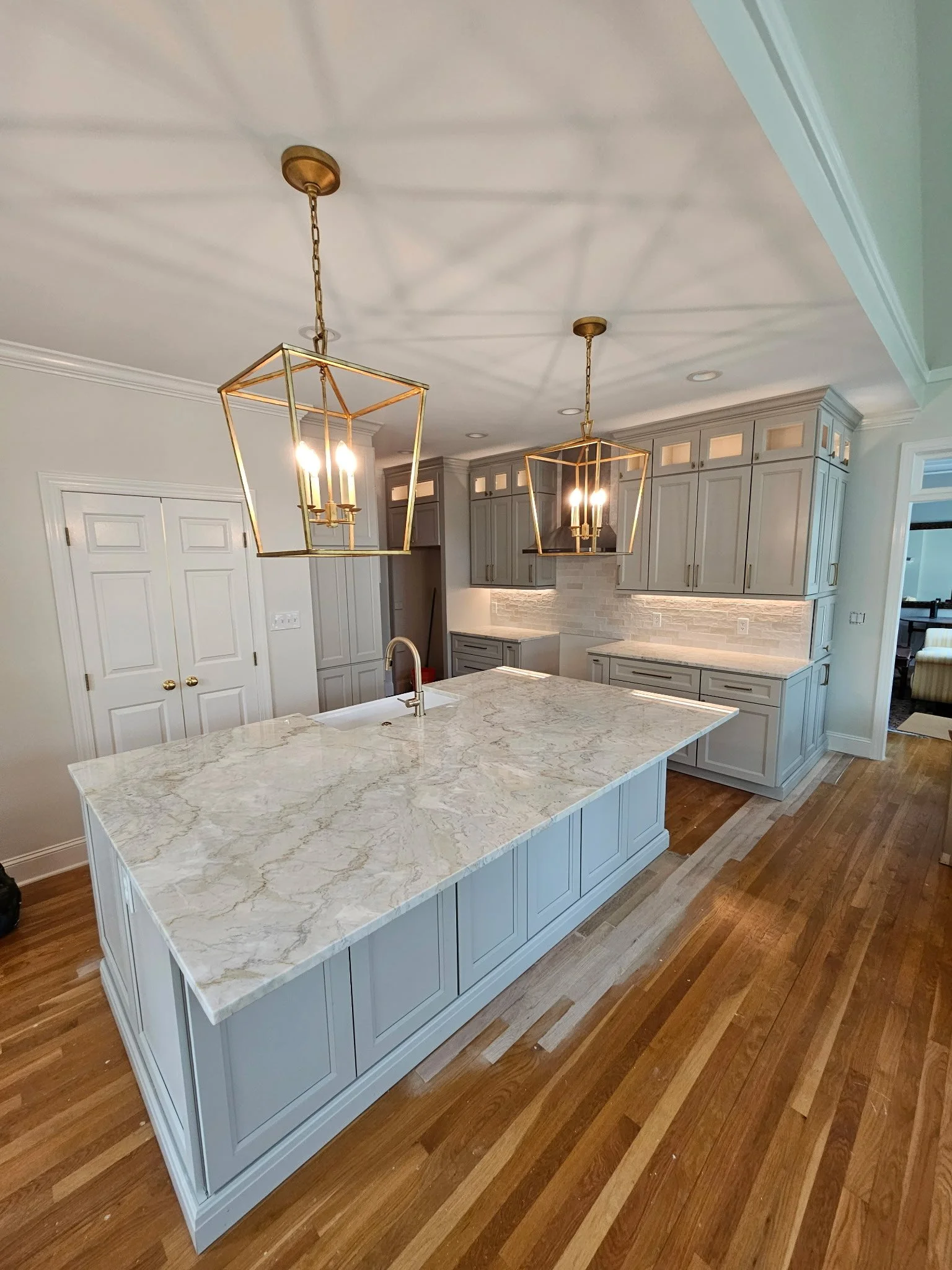 Modern kitchen with white island and light gray cabinets, pendant lights, hardwood floors, and a view into a living room.