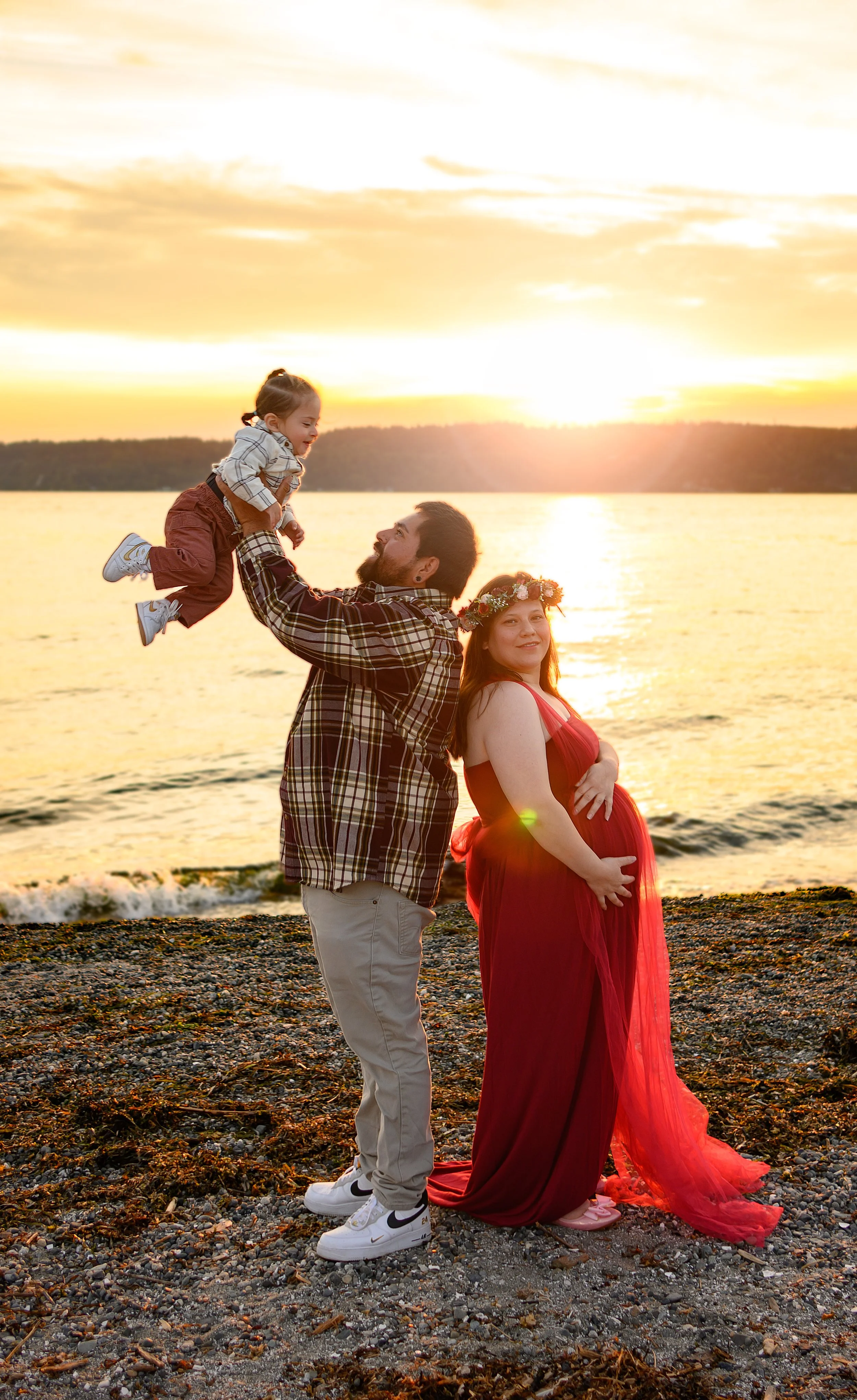 Young woman with red hair wearing a white dress and decorative headband, standing near a body of water during sunset.