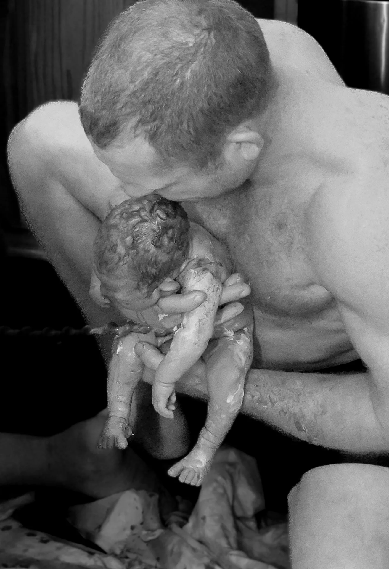 A man with short hair holding a newborn baby in his hands, leaning in to kiss the baby's forehead. The baby is covered in vernix caseosa, the white, cheesy substance that coats newborns. The background appears to contain other babies or similar beings.