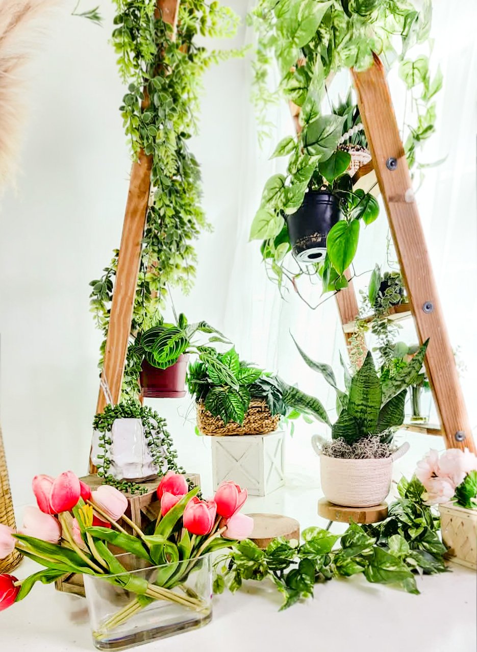 A woman with tattoos and a silver crown smiling with two young girls, who are hugging her and wearing black dresses. They are in front of a floral backdrop with white roses and greenery.