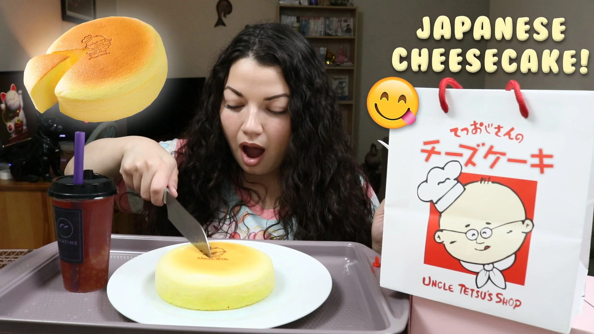 A woman with curly dark hair looks surprised as she cuts into a yellow Japanese cheesecake with a knife. The cheesecake is on a white plate on a tray, and there is a drink with a purple straw next to it. A sign with Japanese characters, a cartoon che