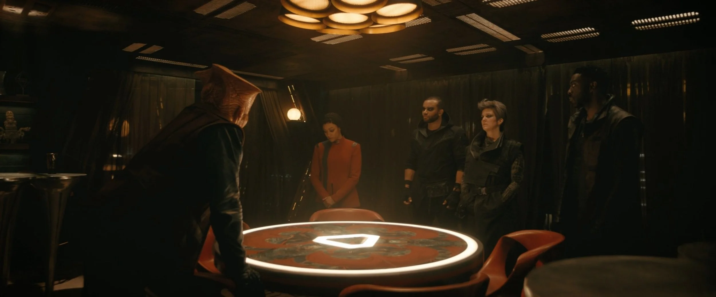 Scene in a dimly lit room with a circular table at the center, featuring the Assassin's Creed logo. Five people stand around the table, one person with a hooded cloak and others in tactical clothing, suggesting a meeting or planning session.