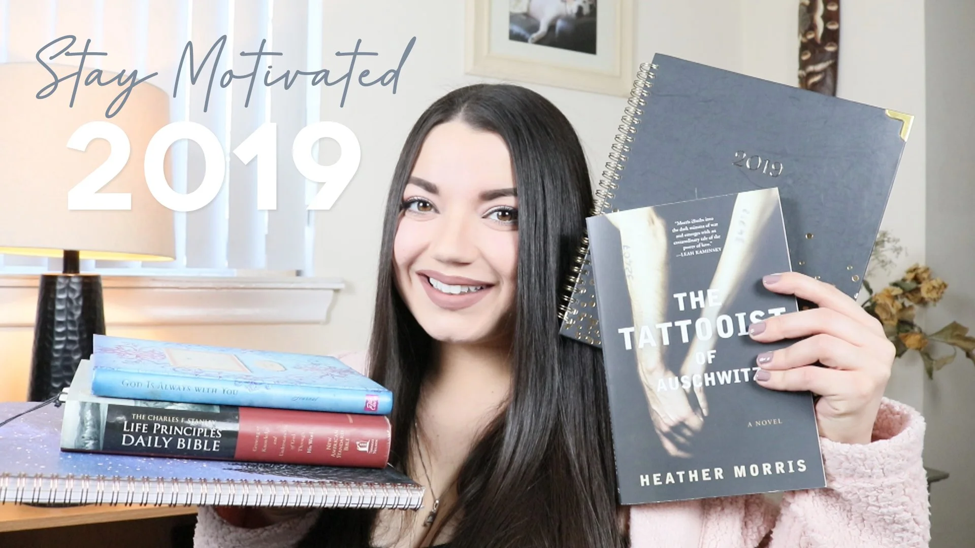 Smiling young woman holding a copy of 'The Tattooist of Auschwitz' by Heather Morris, with a stack of books on a desk including 'God Is Always With You' and 'Life Principles Daily Bible,' in a room with a table lamp, a framed picture, and dried flowe