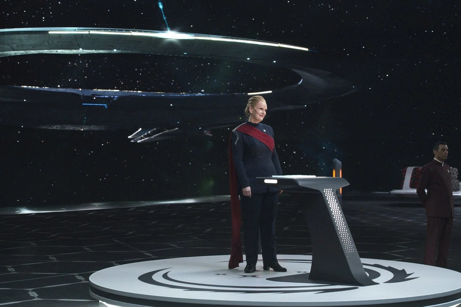 A woman in a dark uniform with red accents stands at a futuristic podium on a spaceship or space station set against a backdrop of stars and a large spaceship in space.