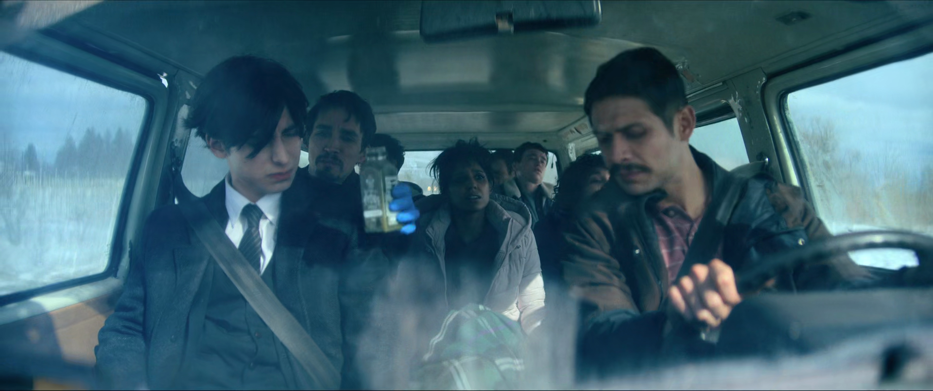 Multiple people sitting in a truck, with the person in the front right driving and older man in the front left holding a small vial, others sitting behind, some looking worried, outside scenery visible through the windows.