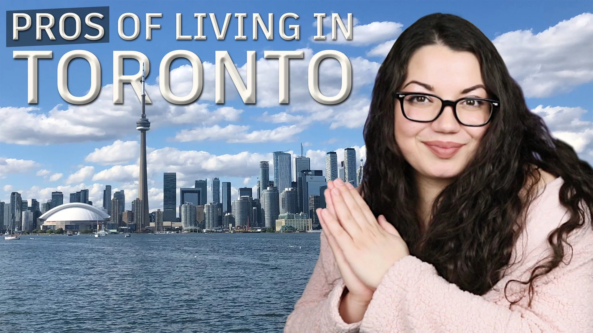A woman with curly dark hair, glasses, and a light pink sweater, smiling with hands pressed together in front of her, with the Toronto skyline and waterfront in the background.