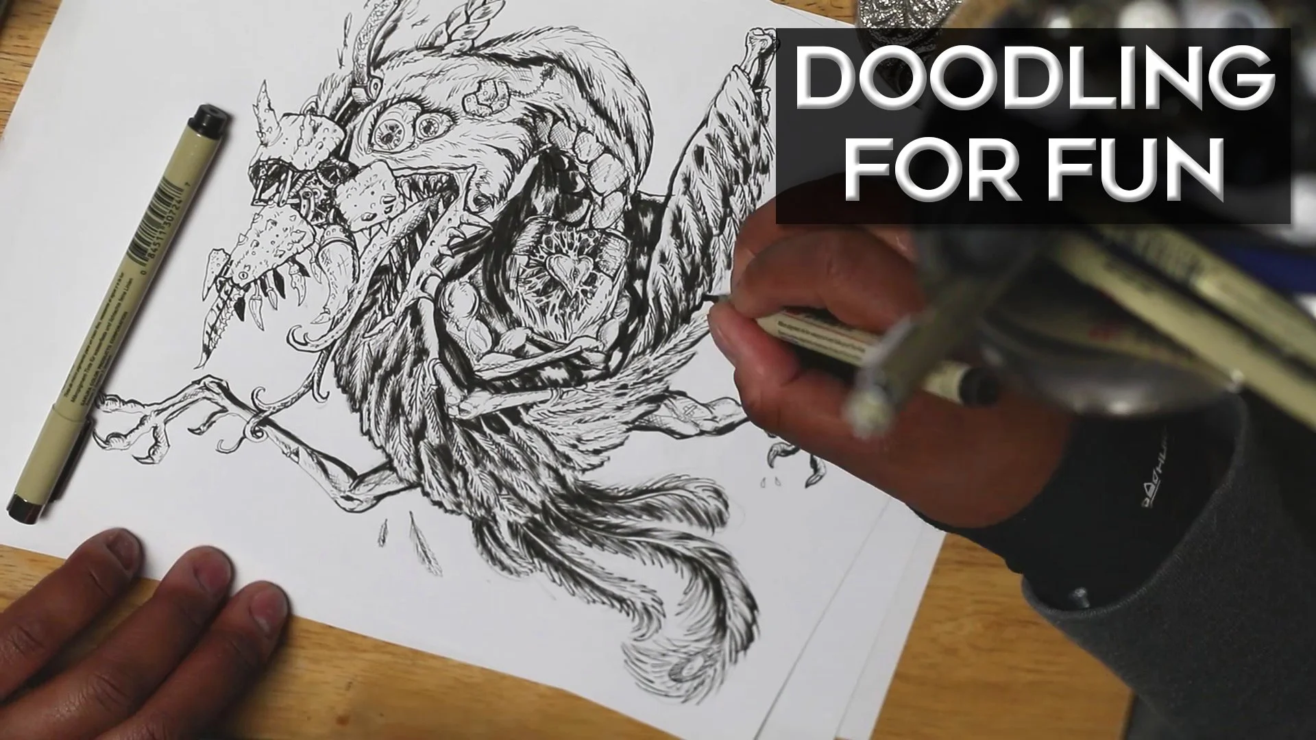 Person drawing a detailed, black-and-white illustration of a creature with a lion's mane, teeth, and claws, on a piece of white paper on a wooden surface, with markers nearby and a hand holding a marker.