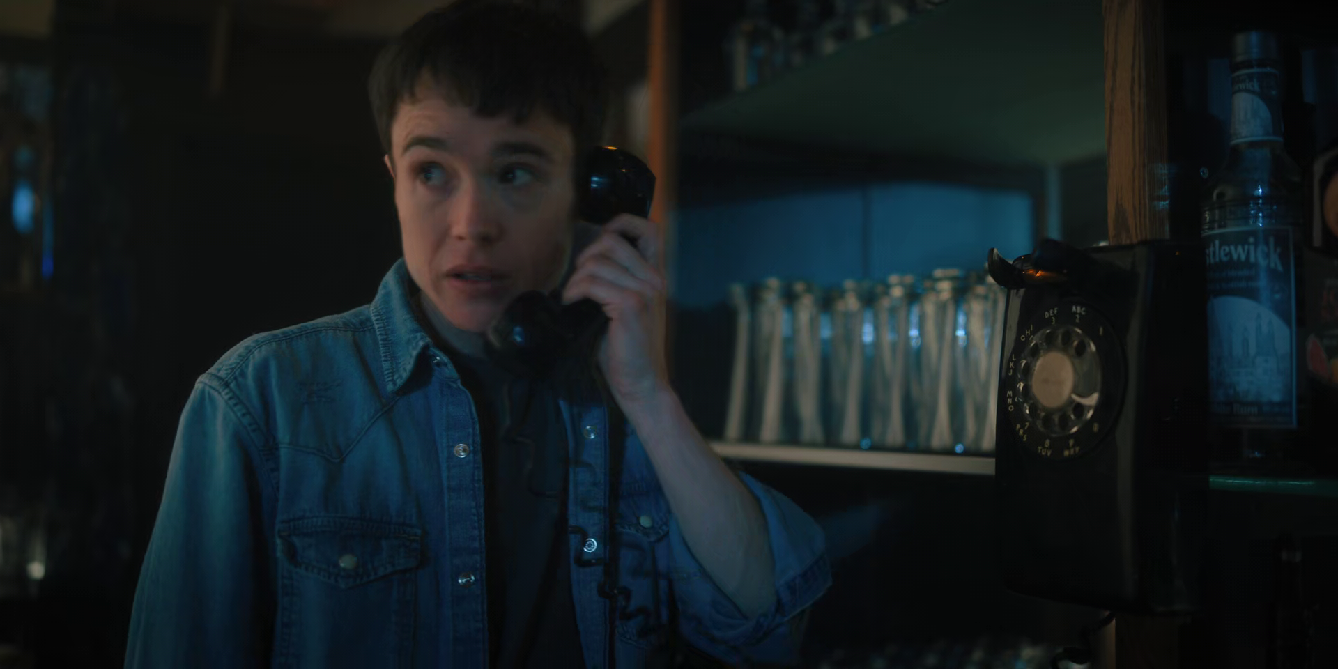 A young man with short dark hair, wearing a denim shirt, using a black rotary phone in a dimly lit bar or pub setting.