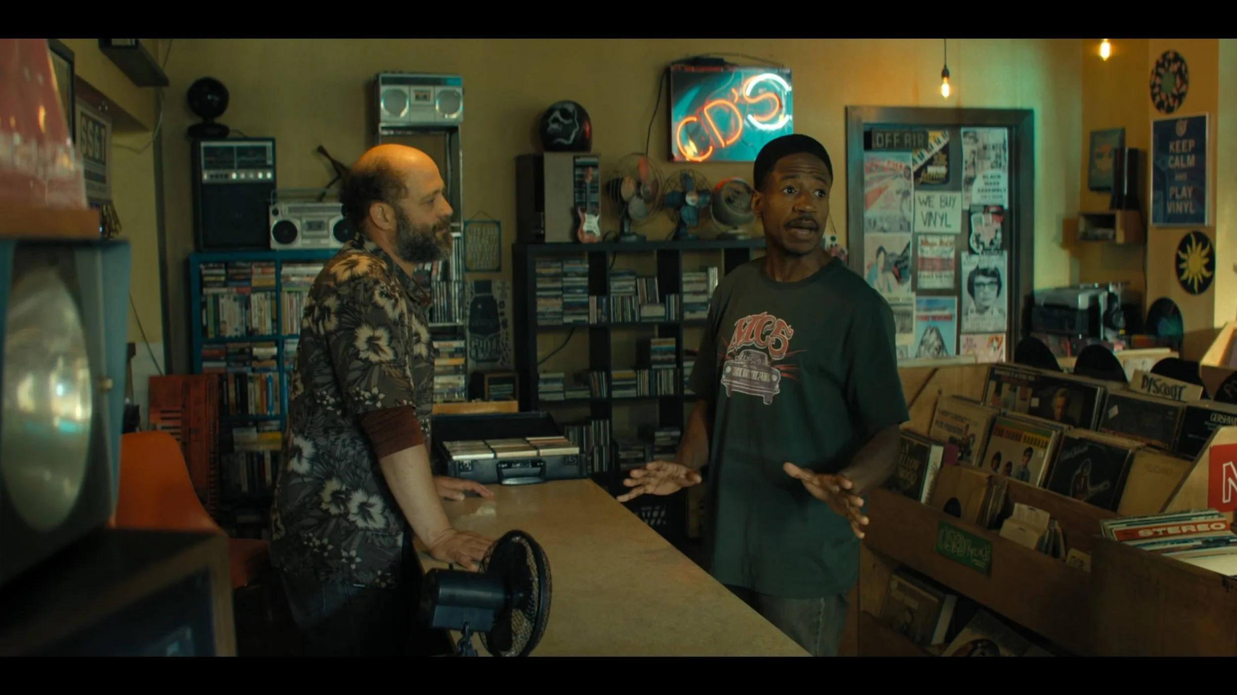 Two men inside a record shop; one wears a Hawaiian shirt and has a beard, the other wears a black T-shirt and is gesturing with his hands, with shelves of records and vinyl albums behind them.