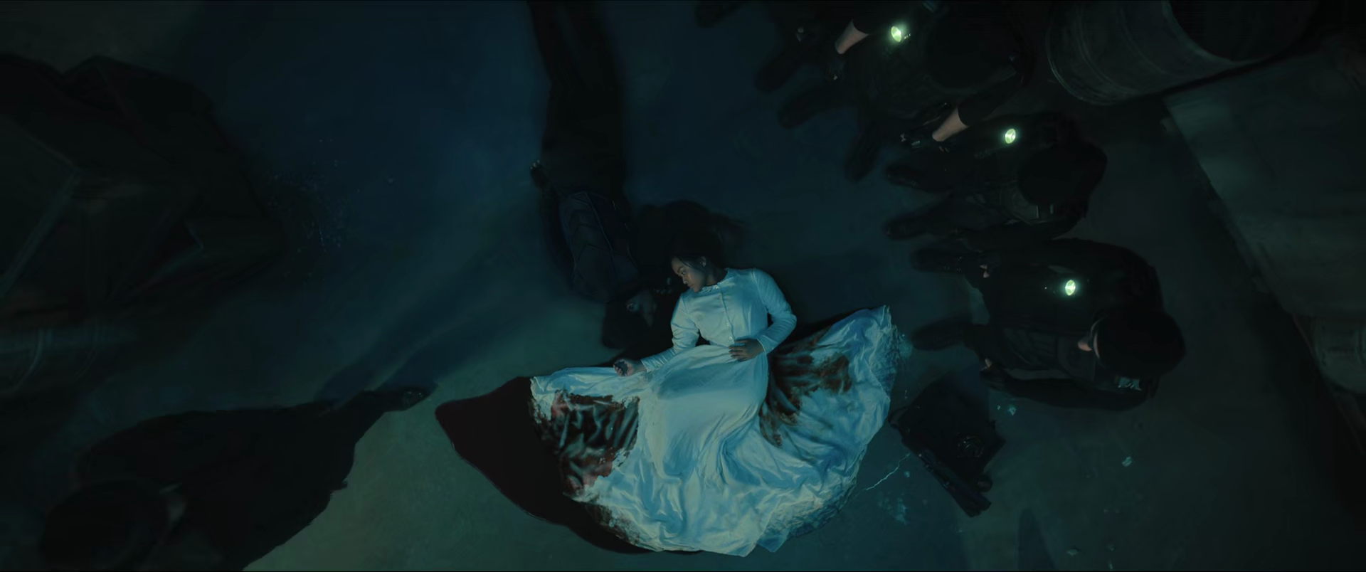 A woman in a white dress lying on the ground surrounded by police officers in dark uniforms with flashlights in a dimly lit setting.