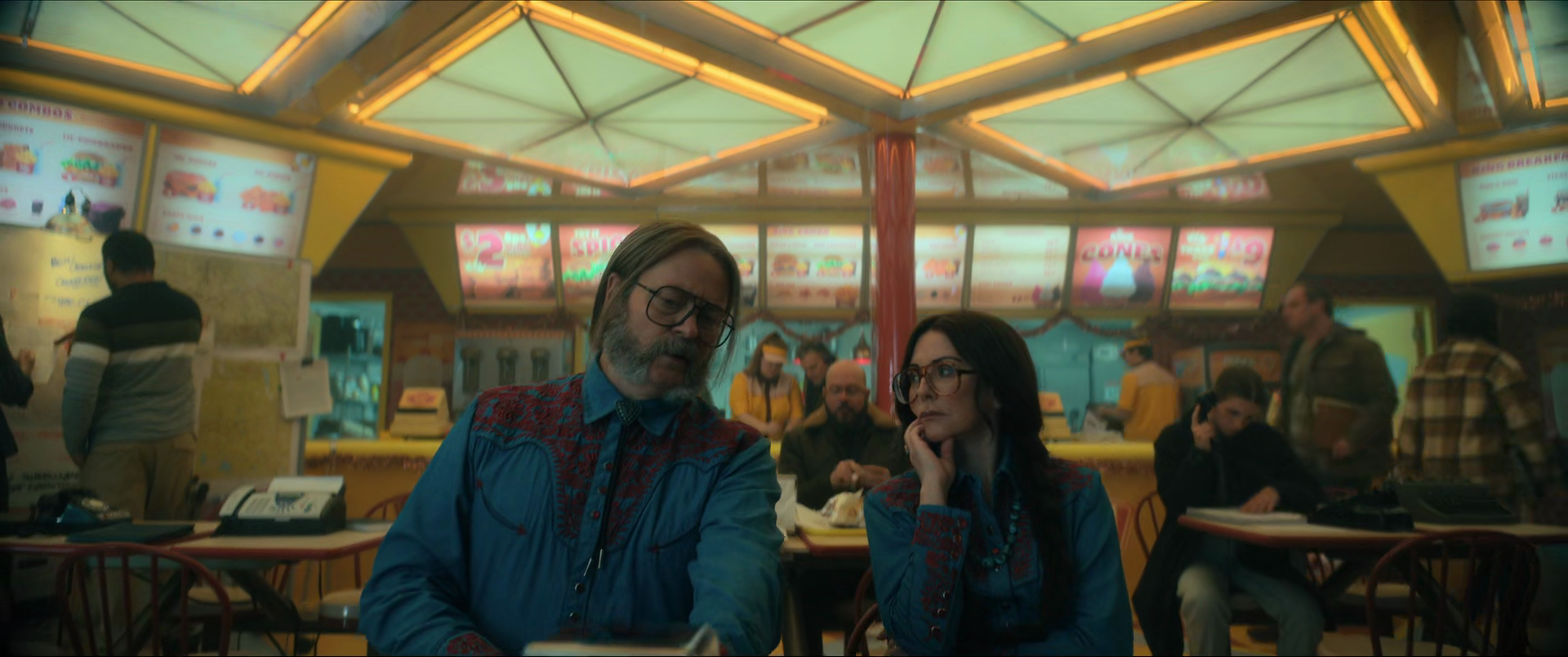 People sitting in a brightly lit fast-food restaurant, two main characters in focus talking, colorful menu signs overhead, others in the background.