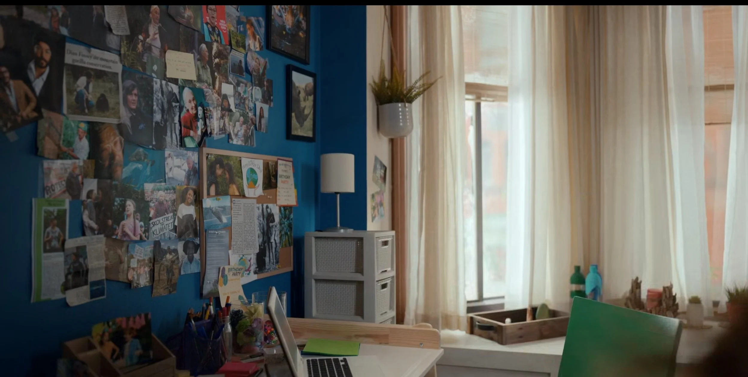 A cozy room with a blue accent wall decorated with many photos and notes, a small desk with a laptop, and a window with sheer curtains letting in natural light.