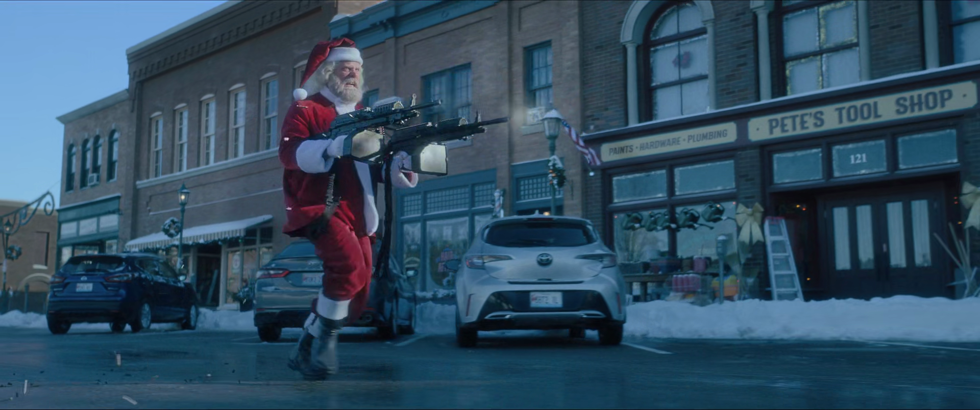 Santa Claus dressed in traditional red and white outfit running down a street, armed with multiple rifles, in a snowy town setting.