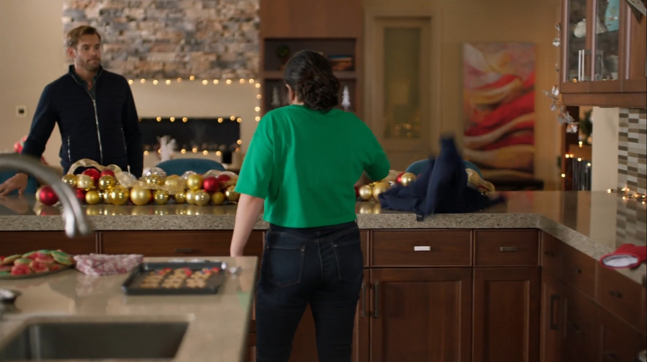 A woman with dark hair wearing a green shirt and jeans standing in a kitchen, decorated with Christmas ornaments, with a man in a dark jacket in the background near a fireplace with holiday lights.