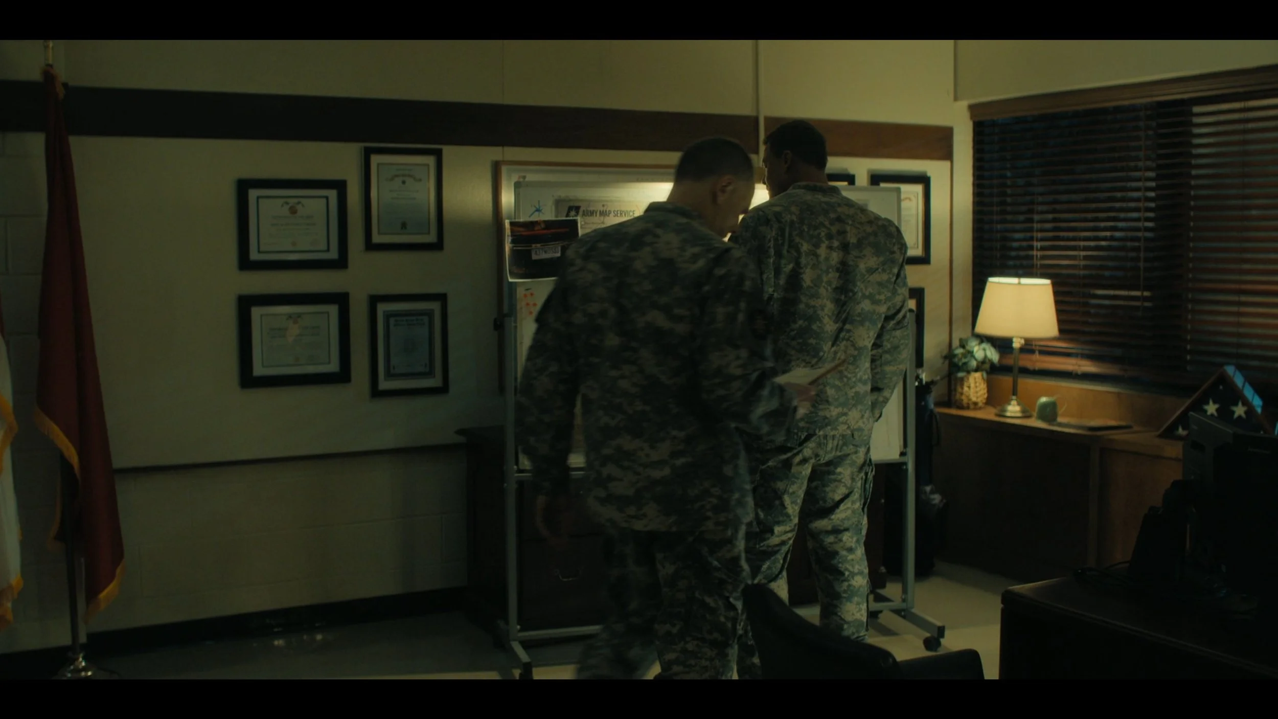 Two soldiers in camouflage uniforms standing in a dimly lit room, looking at a whiteboard with documents or maps, with framed certificates on the wall and a lamp on a wooden table near a window with blinds.
