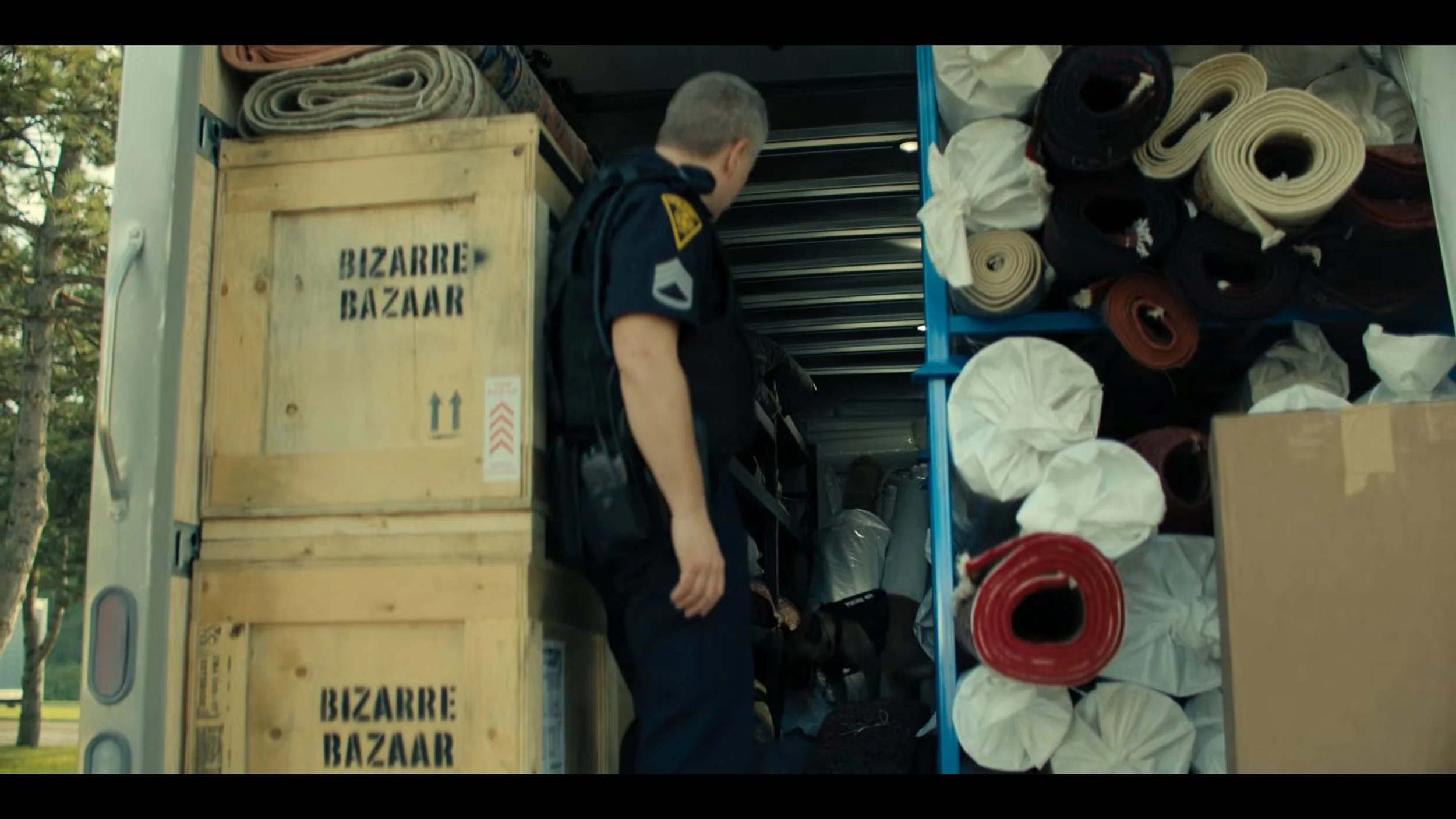 A person in a police uniform with a backpack looking inside a truck filled with rolled-up mats and boxes labeled 'Bizarre Bazaar'.