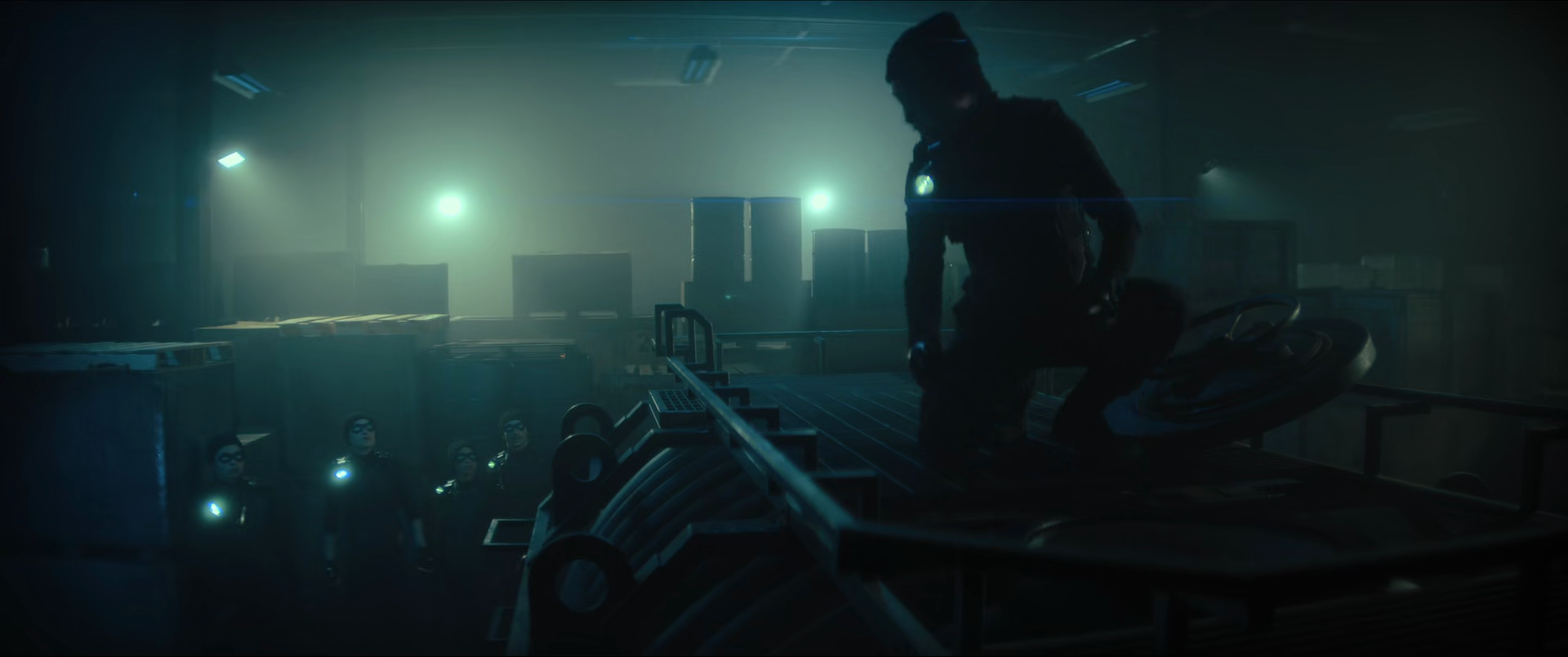 Silhouette of a person in a dark room with a bright light behind, surrounded by several smaller figures wearing masks and glowing accents, as if in a futuristic or cinematic setting.