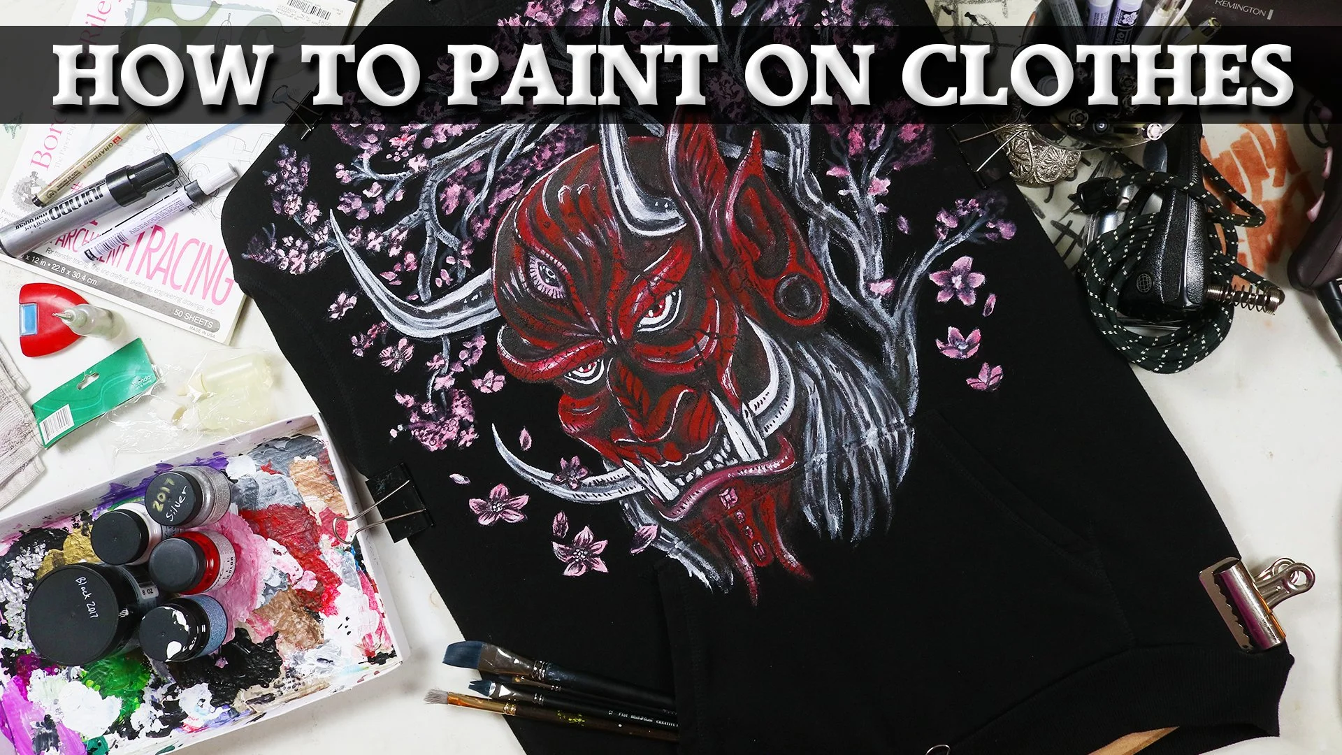 A clothing item with a painted tiger face design surrounded by pink cherry blossoms, placed on a cluttered artist's workspace with paint bottles, brushes, and art supplies.